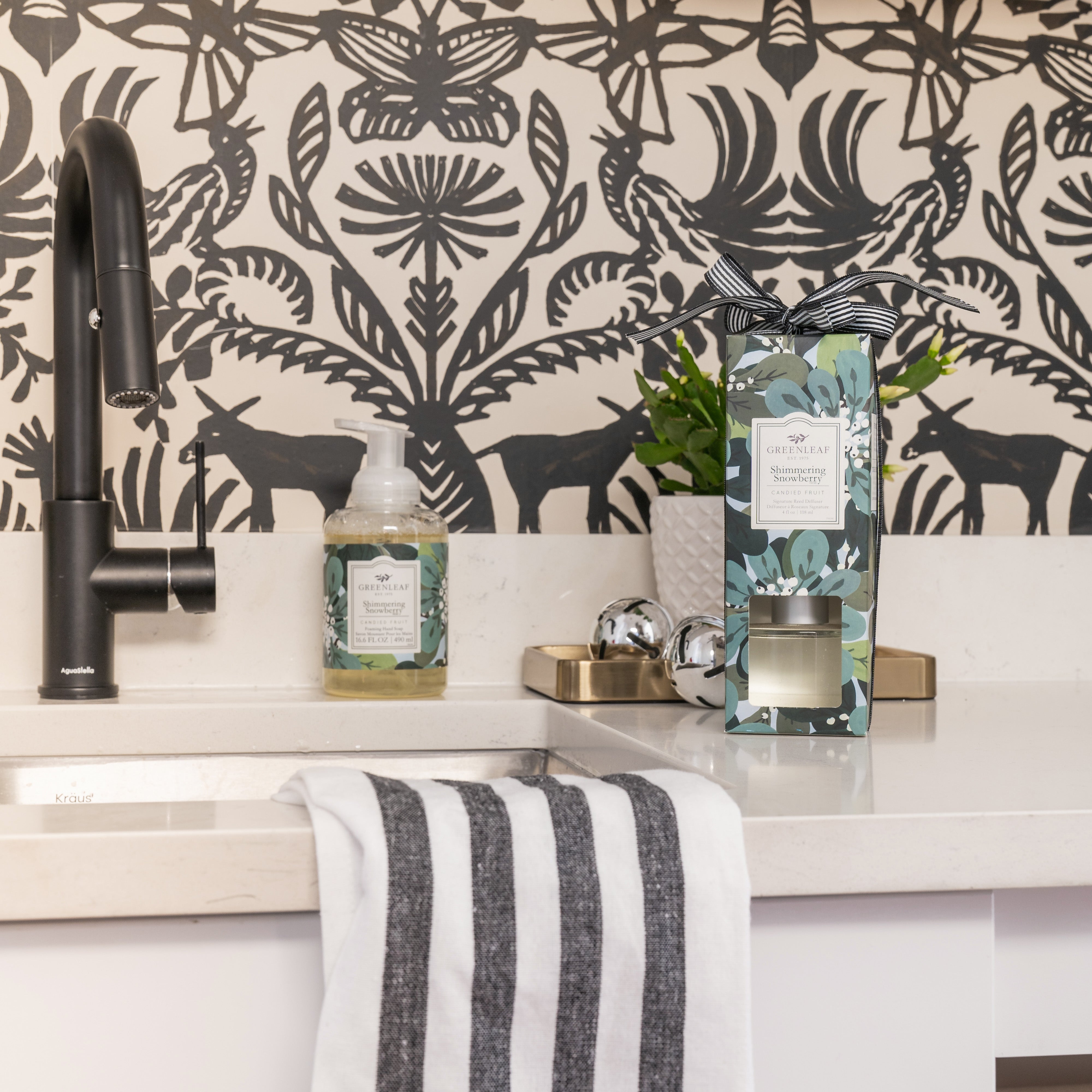 A bathroom sink area with a black faucet, striped towel, soap dispenser, decorative box, plant, and the elegant Greenleaf Gifts Shimmering Snowberry Reed Diffuser. The background features black and white floral and bird patterned wallpaper.