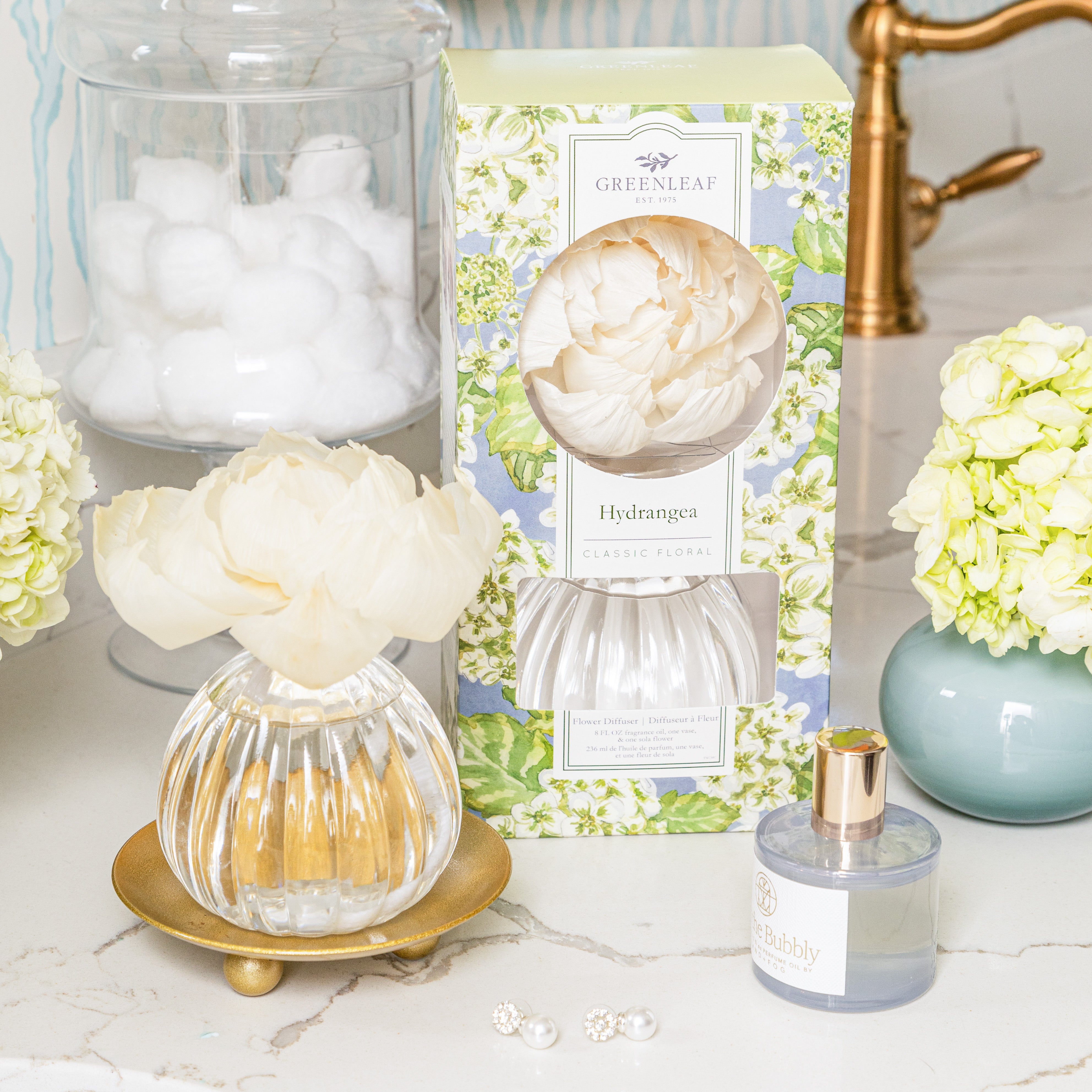 A Greenleaf Gifts Hydrangea Flower Diffuser sits on a gold tray near a Greenleaf Hydrangea fragrance box, a Bubbly-labeled bottle, and white cotton balls on a marble bathroom counter with hydrangea flowers.
