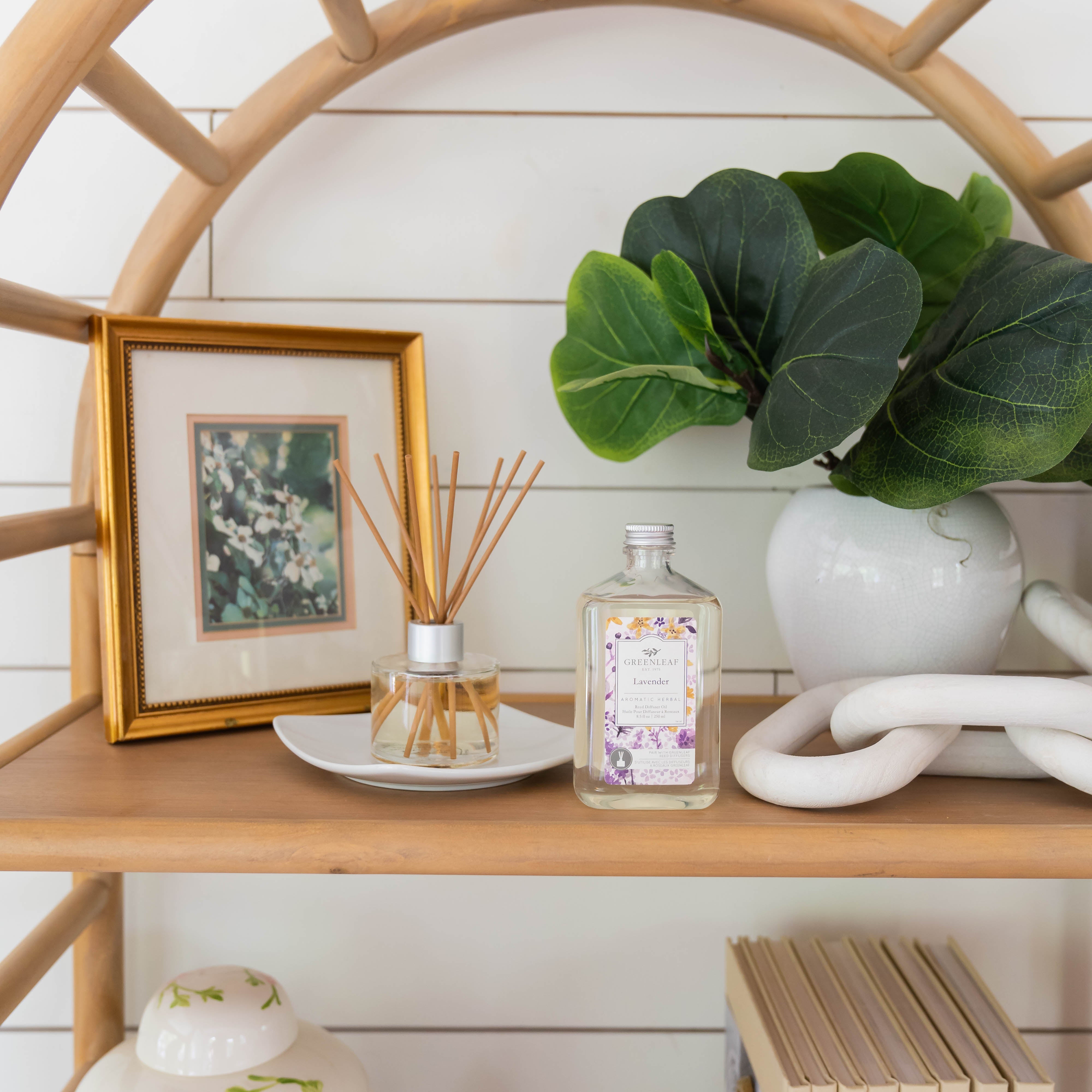 A wooden shelf displays a framed floral print, a Greenleaf Gifts Lavender Diffuser Oil Refill, a Greenleaf Reed Diffuser with lavender scent, a white vase with green leaves, white decorative links, and a jar with a green pattern.