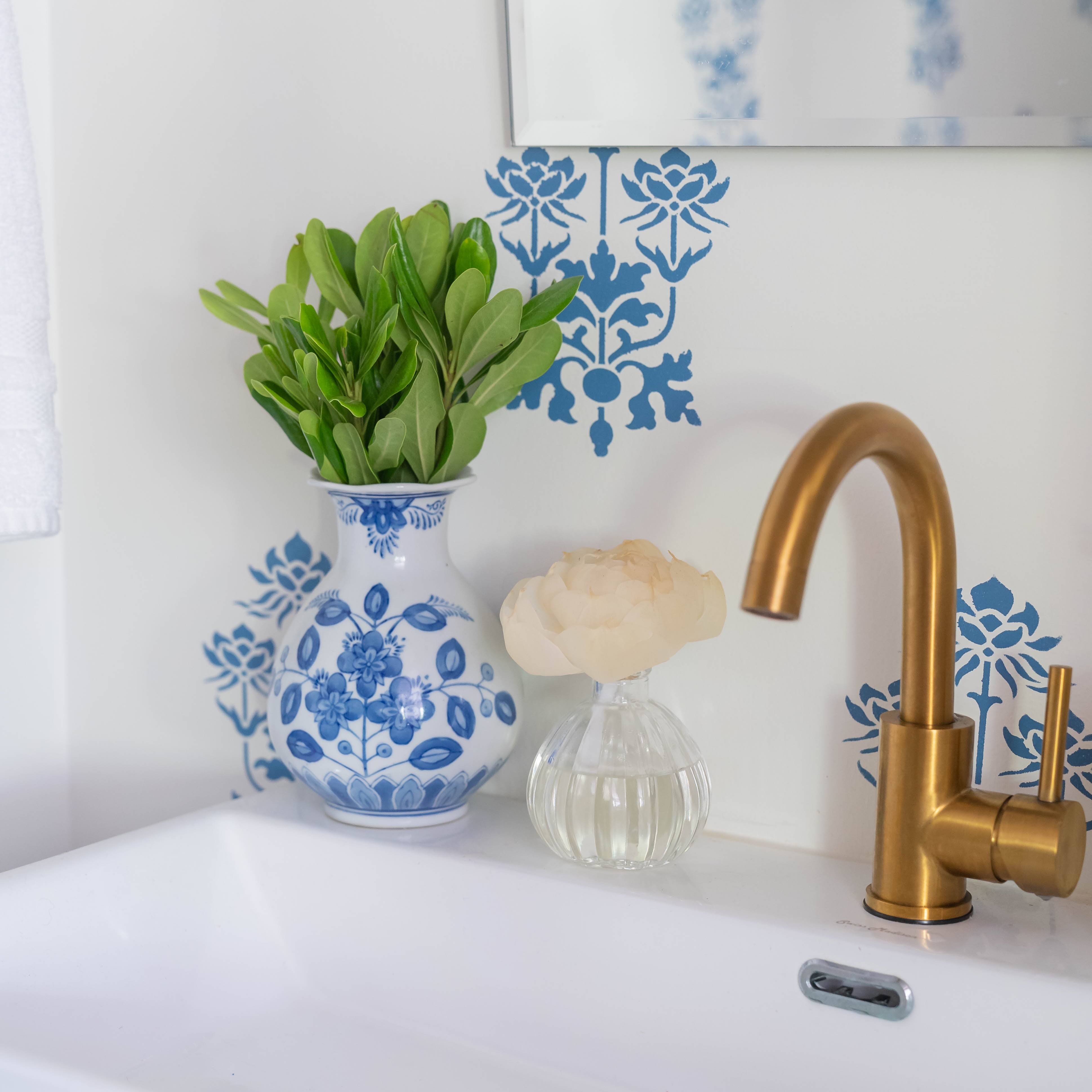 A white sink with a gold faucet, a Greenleaf Gifts Flower Diffuser-Classic Linen in an 8 oz blue and white vase with green leaves, a small glass vase with a white flower, and blue floral patterns on the wall behind them.