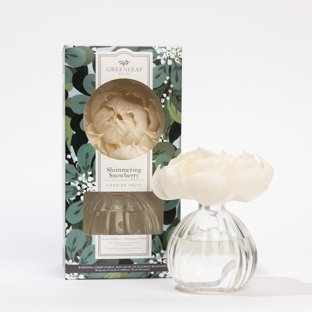 The Greenleaf Gifts Shimmering Snowberry Flower Diffuser features a clear round vase with fragrance oil and a white sola paper flower, displayed next to its decorative green and floral-patterned box.