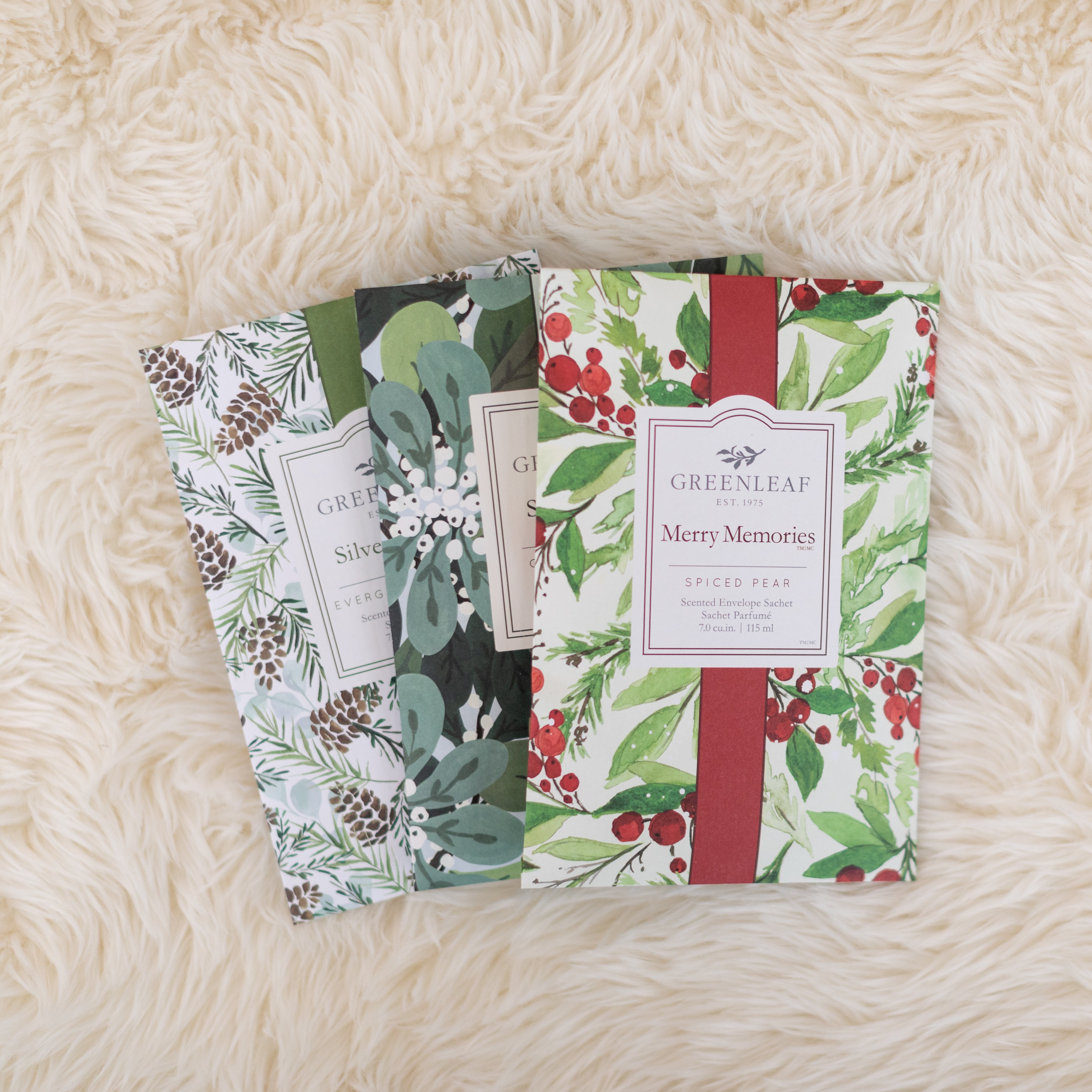 Two Greenleaf Gifts Merry Memories Sachets with botanical designs rest on a cream fluffy surface—one shows pine and berries with a red Merry Memories band, the other features green foliage and a white band.
