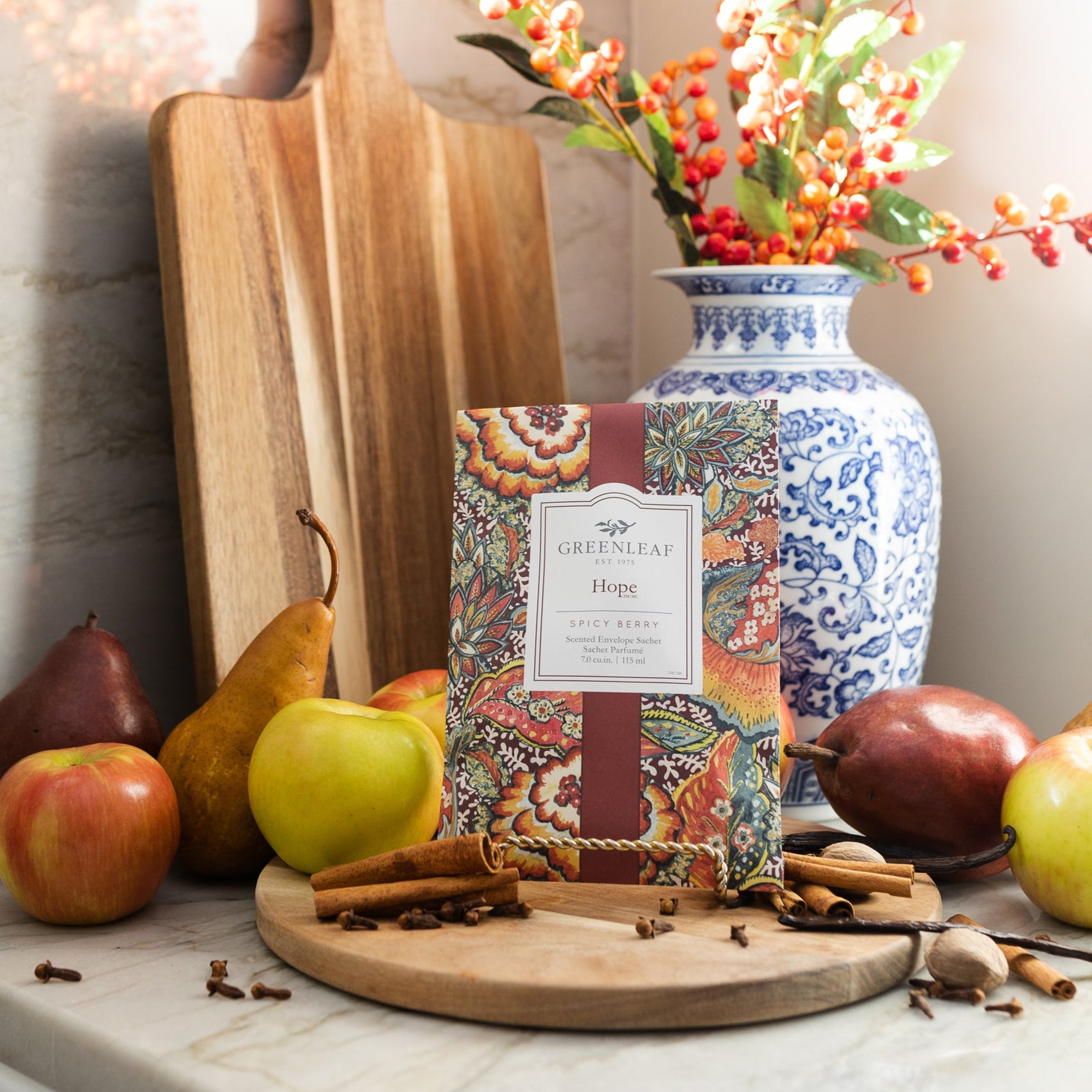 A decorative kitchen scene features apples, pears, spices, and a patterned Greenleaf Gifts Hope Sachet for home fragrance on a round wooden board, beside a blue-and-white vase with orange berries and cutting boards as sunlight streams in.