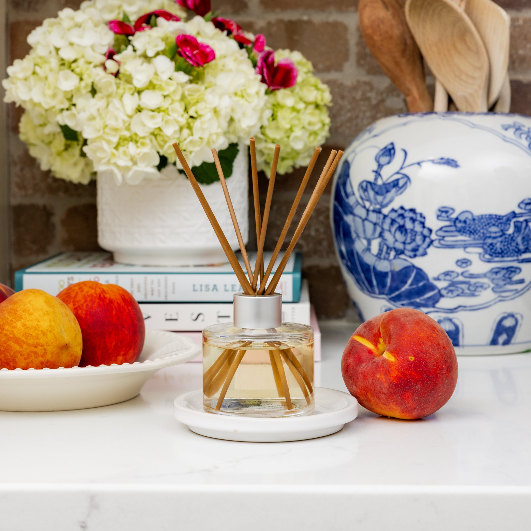 The Greenleaf Gifts Coco Peach Reed Diffuser with fiber reeds sits on a white plate beside peaches, a fruit bowl, white flowers in a vase, a blue-and-white utensil jar, and stacked books on a white countertop.
