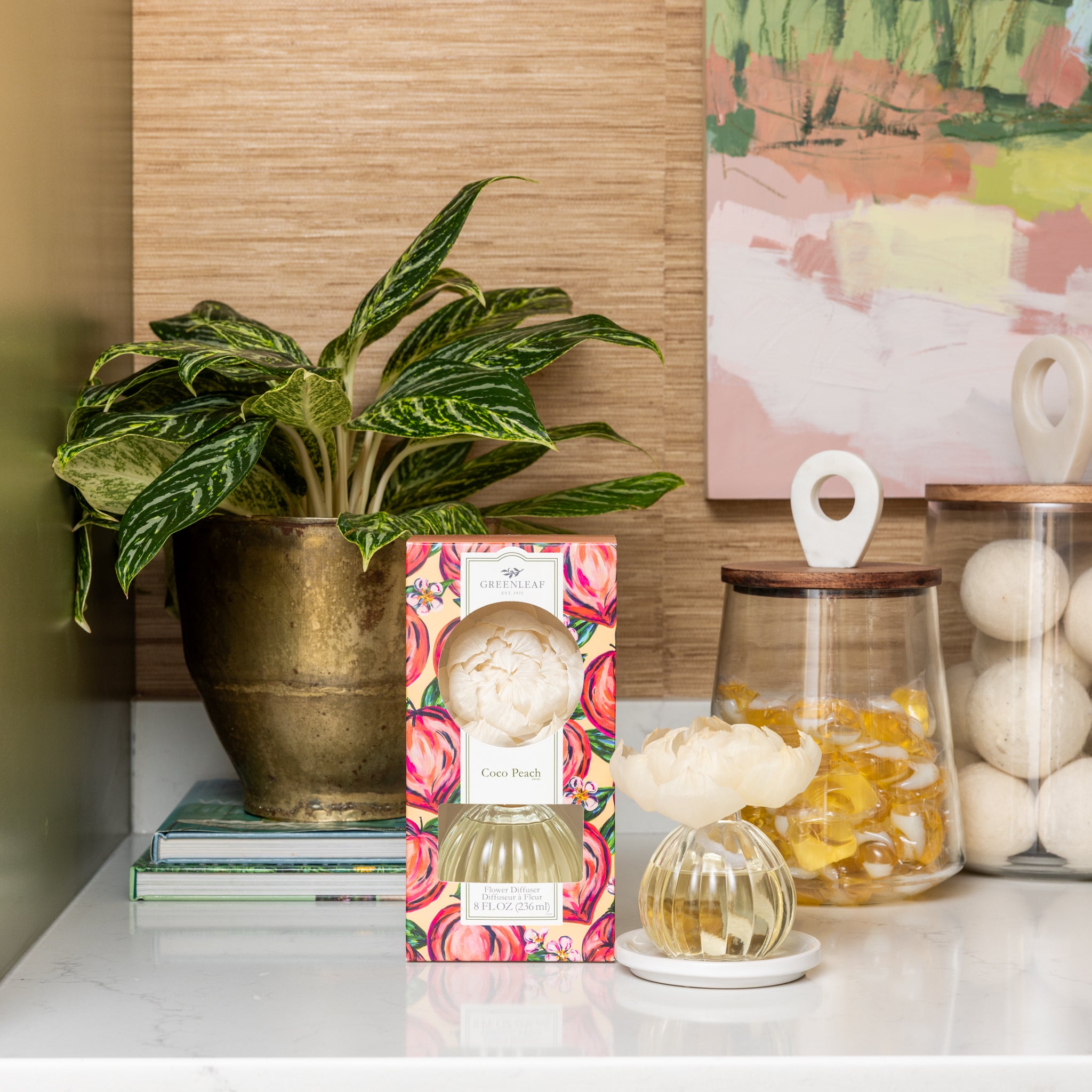 A decorative countertop features a potted plant, a Greenleaf Gifts Coco Peach Flower Diffuser with sola paper flower, glass jars containing yellow capsules and white balls, stacked books, and abstract wall art in the background.
