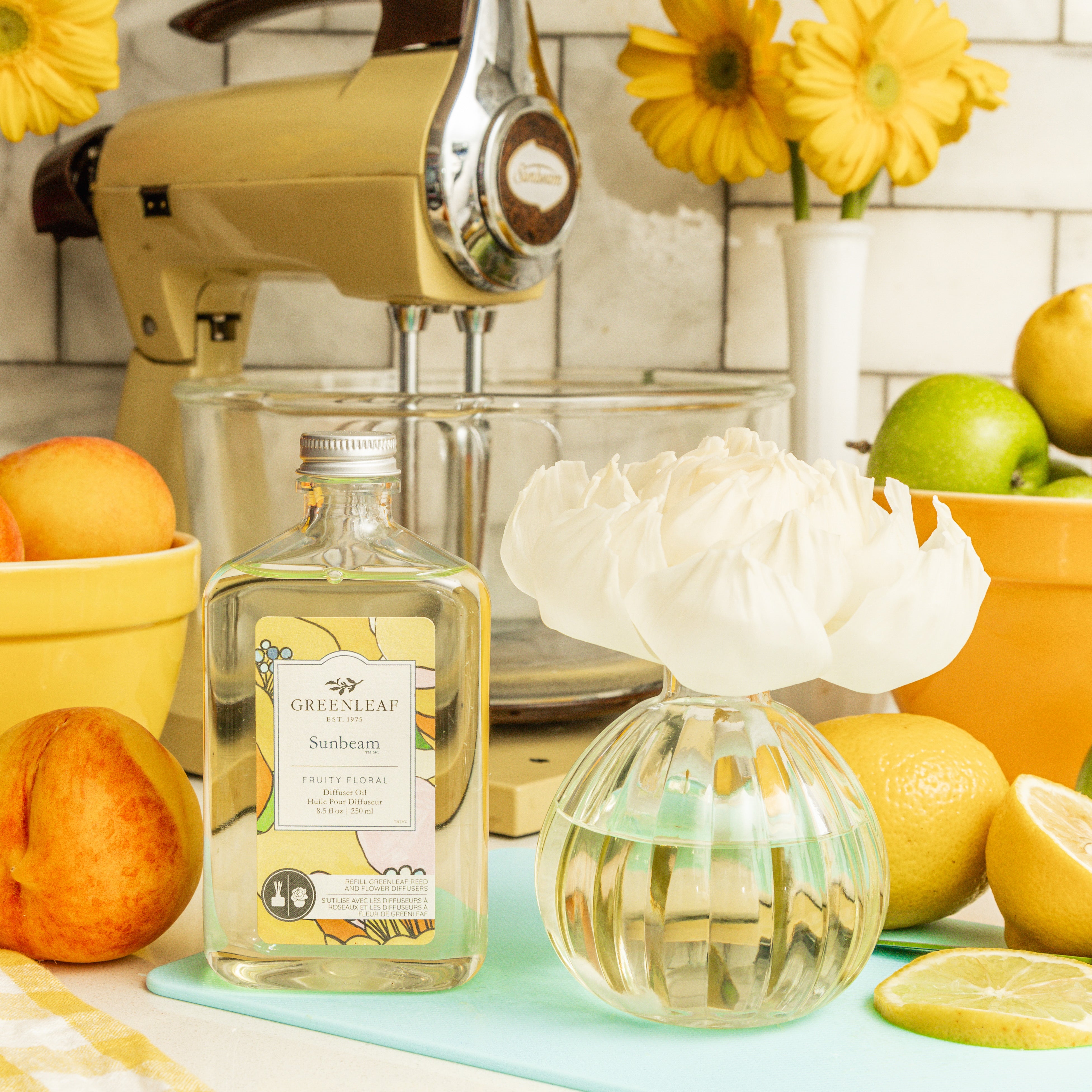 A Greenleaf Gifts Sunbeam Diffuser Refill Oil bottle stands beside a glass vase with a white flower, surrounded by fresh peaches, lime, lemon, yellow bowls, flowers, and a vintage mixer on the kitchen counter.