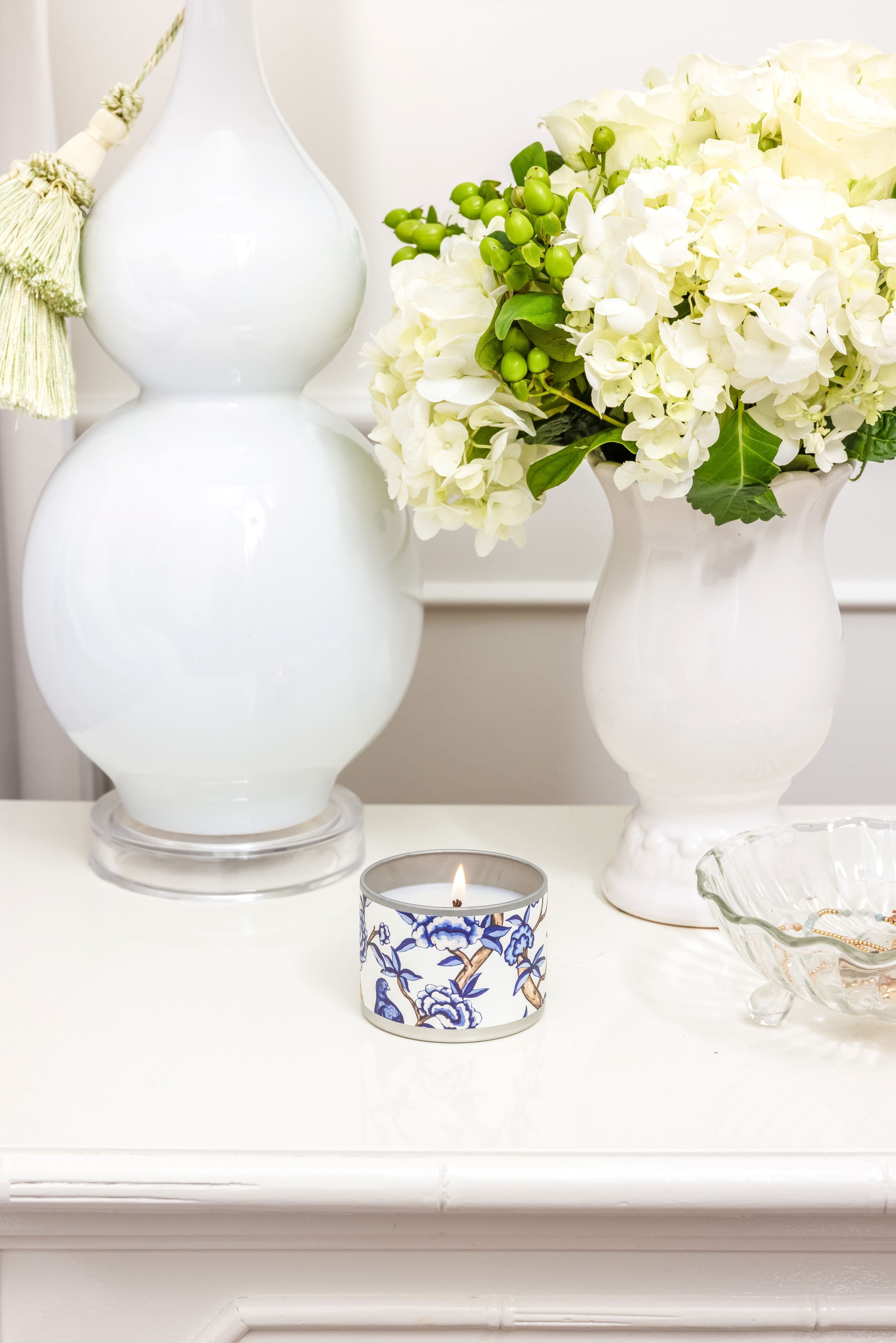 The Greenleaf Gifts Classic Linen Tin Candle sits on a white surface next to a white lamp, a vase of hydrangeas with green berries, and a clear glass dish.