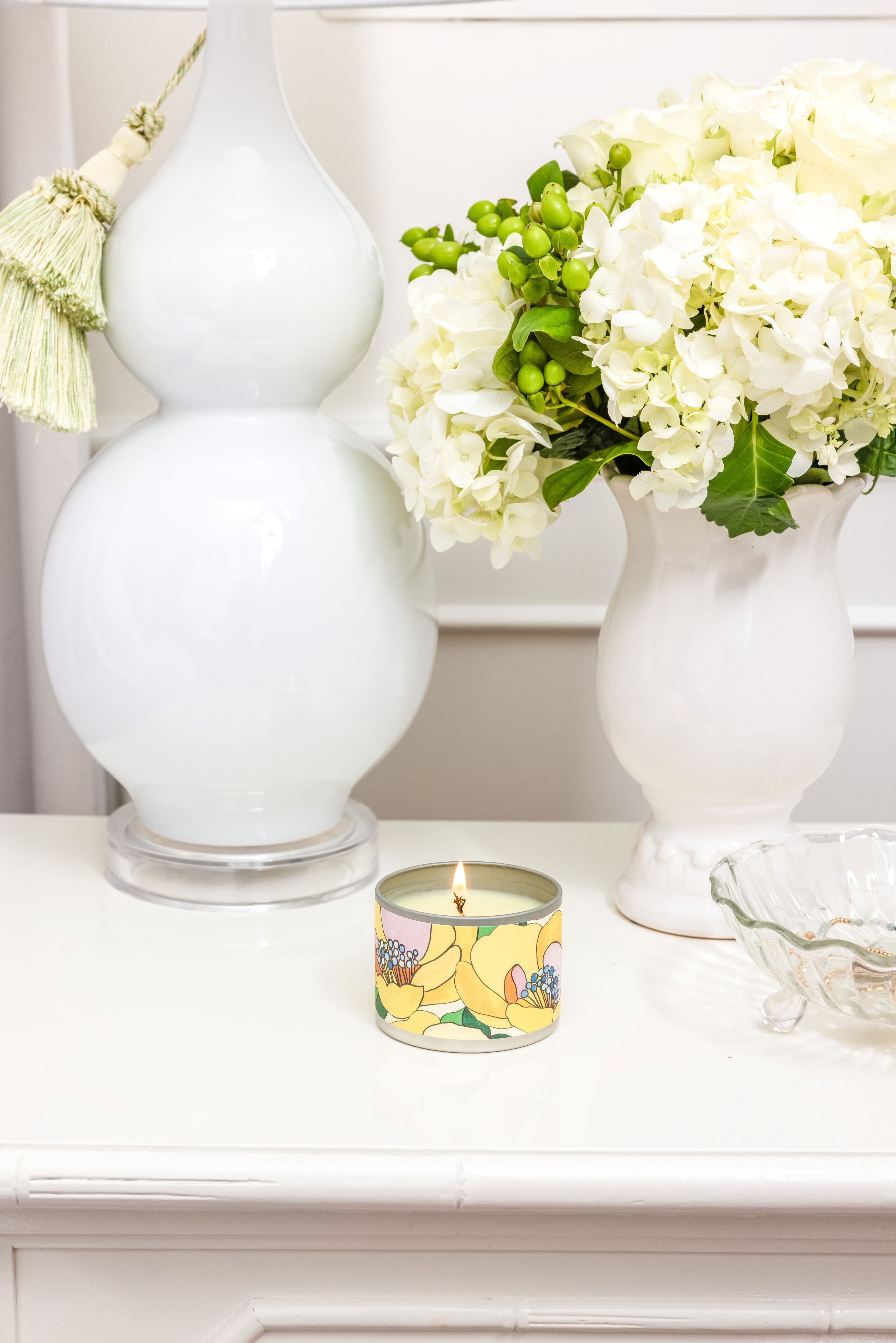 The Sunbeam Tin Candle by Greenleaf Gifts sits on a white table beside a vase of hydrangeas and green berries, a large white lamp with a tassel, and a clear glass dish.