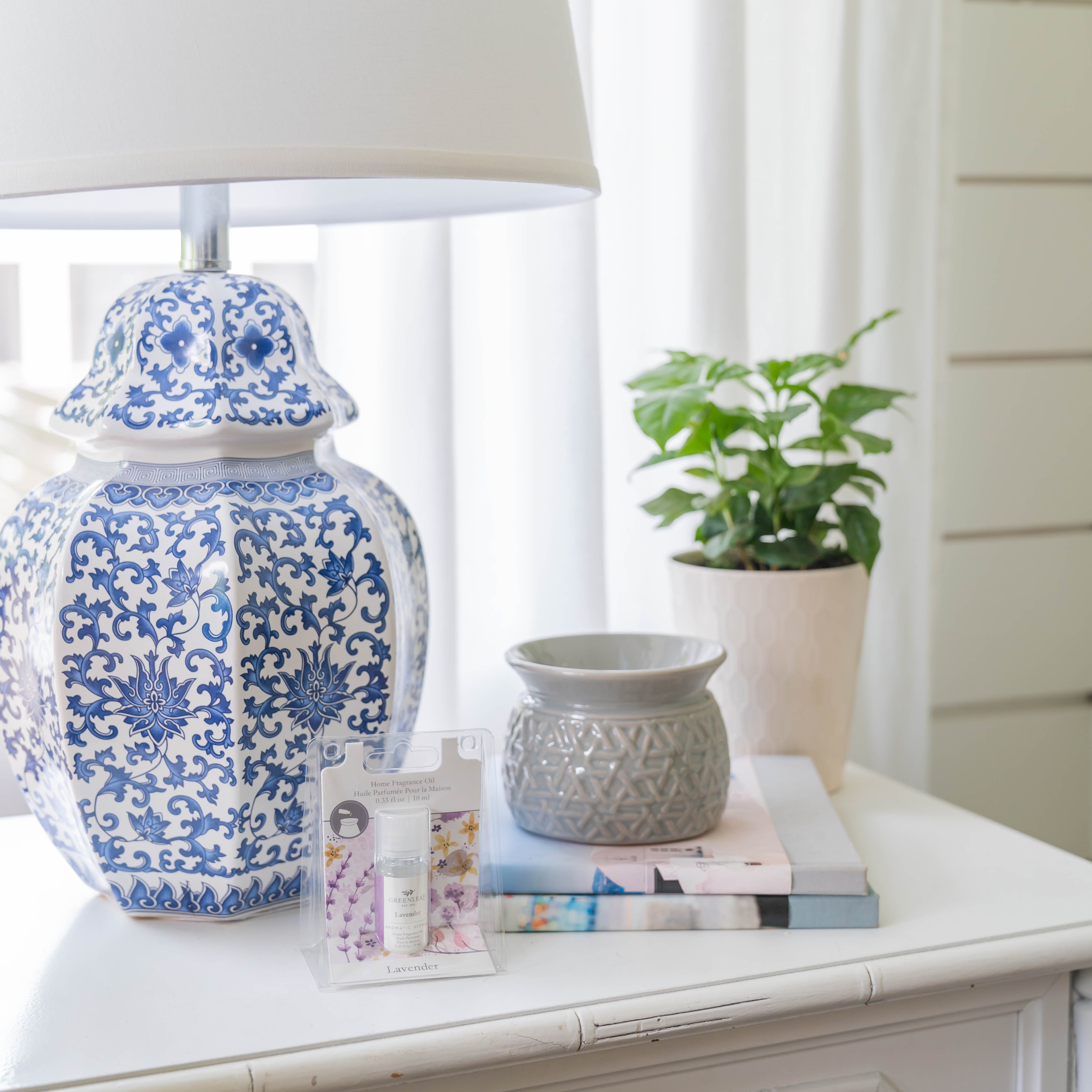 A blue and white patterned lamp, a small potted plant, a gray textured vase, stacked books, and Greenleaf Gifts Lavender Home Fragrance Oil are neatly arranged on a white bedside table.