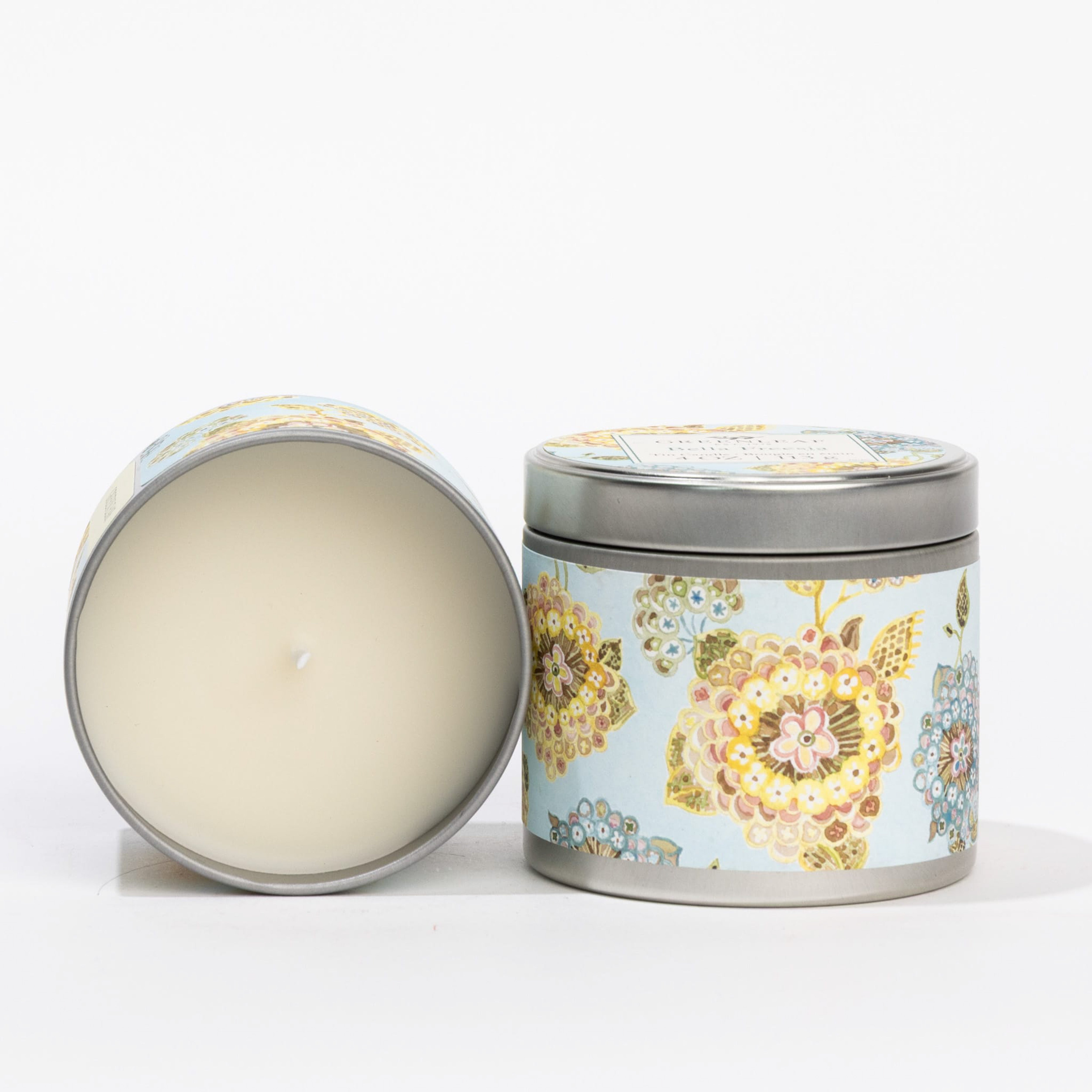 Two Greenleaf Gifts Bella Freesia Tin Candles are displayed against a plain white background—one upright with its lid on, and the other lying on its side to reveal the white wax and delicate floral tin design.