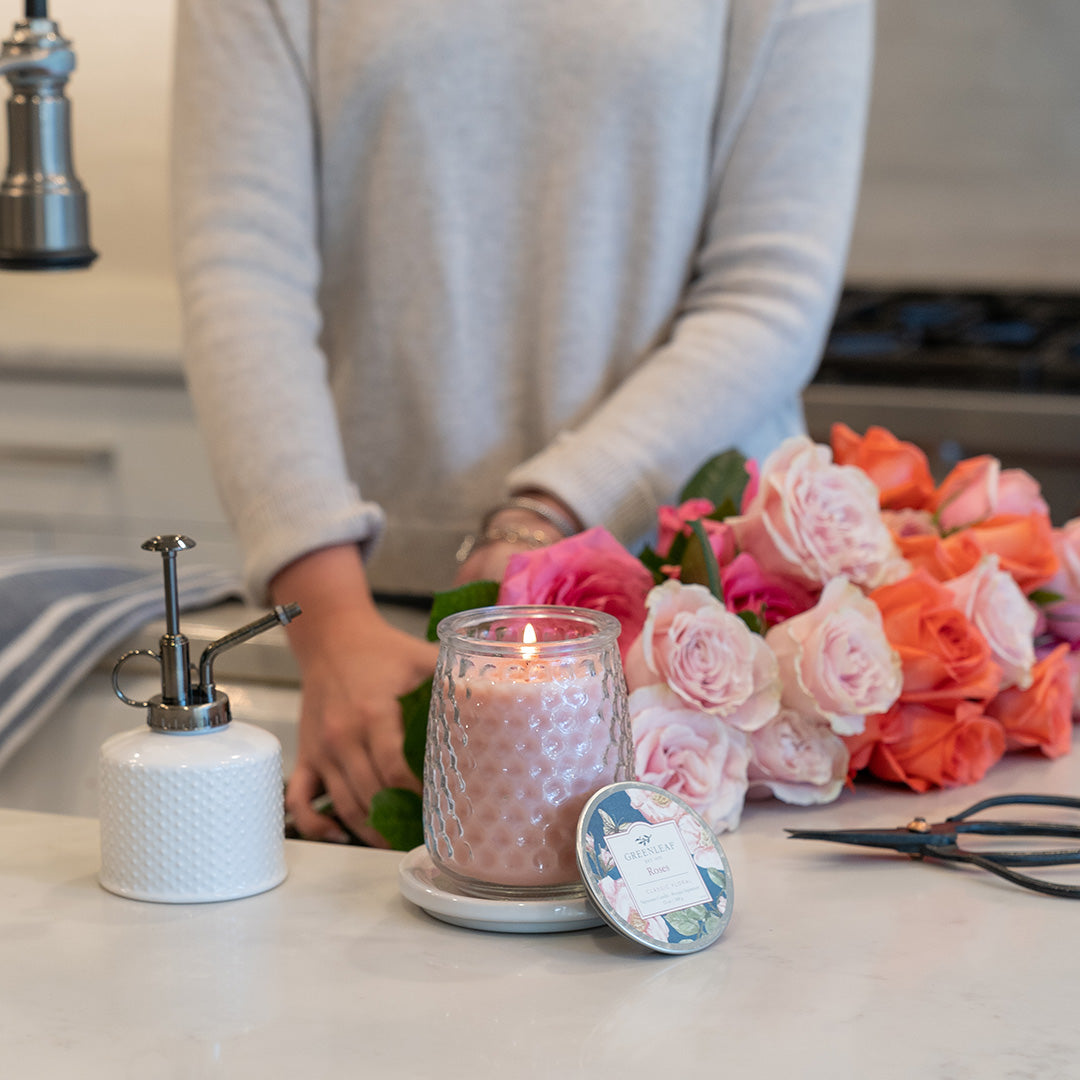 A person in a light sweater stands at a kitchen counter with a lit Greenleaf Gifts Roses Signature Candle, a bouquet of pink and orange roses, a soap dispenser, and scissors nearby.