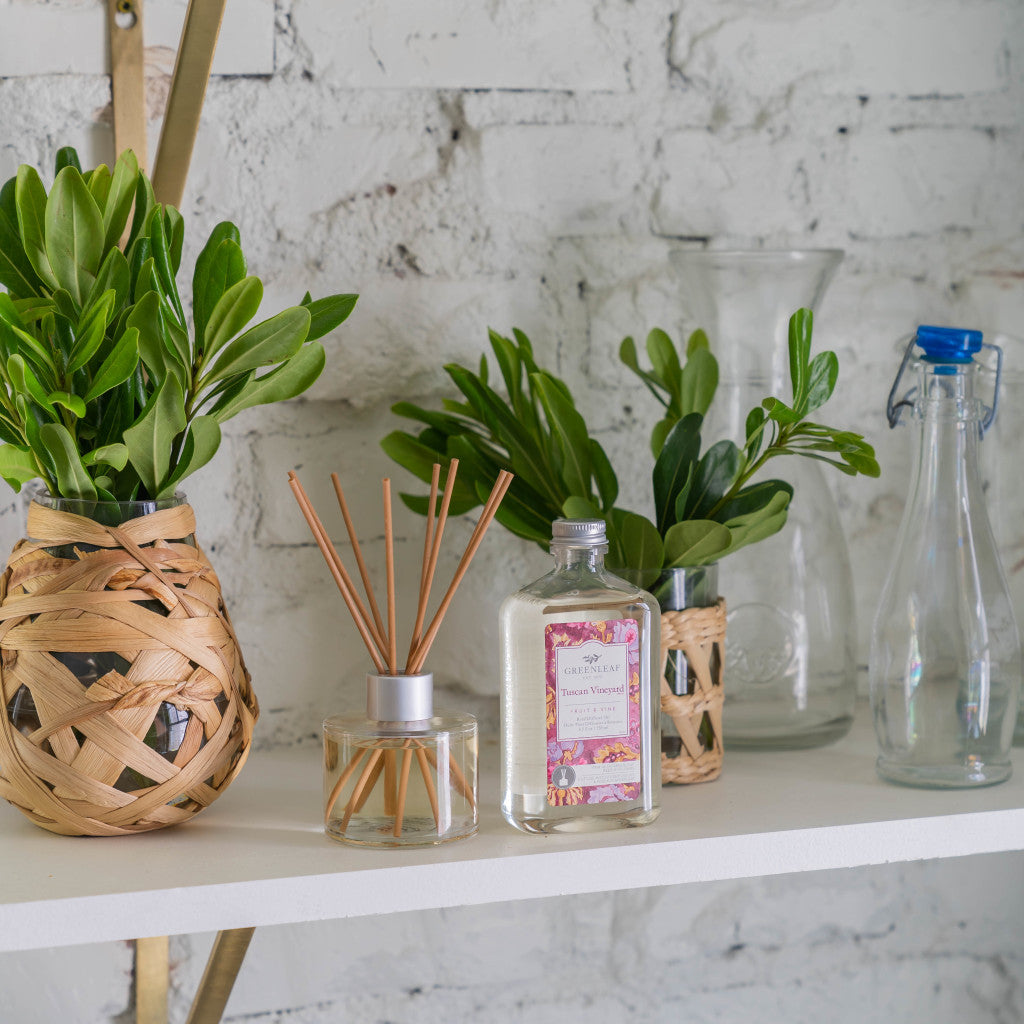 A white shelf displays two glass vases with green leaves in woven baskets, a Greenleaf Gifts Tuscan Vineyard Diffuser Oil Refill, a bottle with a decorative label, a clear carafe, and a glass bottle with a blue cap against a white brick wall.