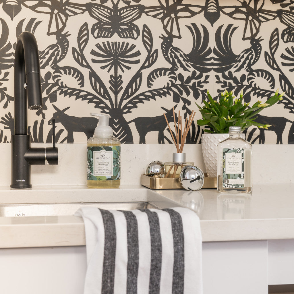A modern kitchen sink area with a black faucet, striped towel, soap dispenser, decorative tray featuring Greenleaf Gifts’ Shimmering Snowberry Diffuser Oil Refill, hand soap, potted plant, and a black-and-white nature-themed backsplash.