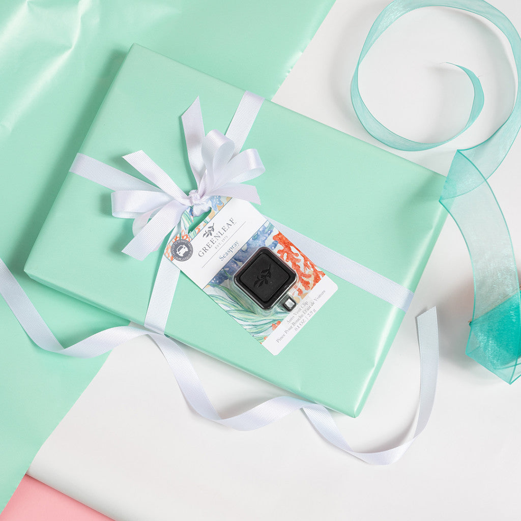 A gift wrapped in mint green paper with a white ribbon, topped with a Greenleaf Gifts Seaspray Auto Vent Clip car air freshener and a colorful tag, surrounded by white and teal sheer ribbon on a white background.