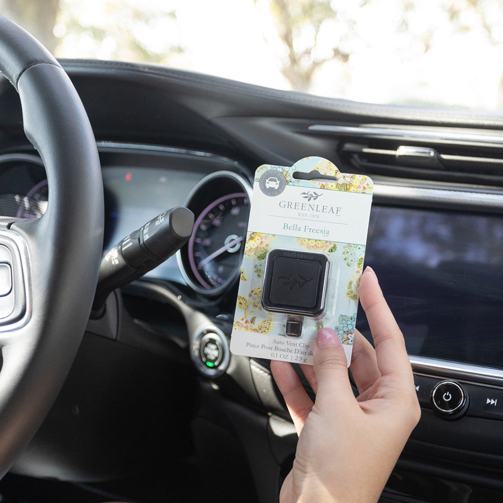 A person holds a Greenleaf Gifts Bella Freesia Auto Vent Clip in its packaging inside a car, with the steering wheel and dashboard in view, highlighting this signature fragrance air freshener.