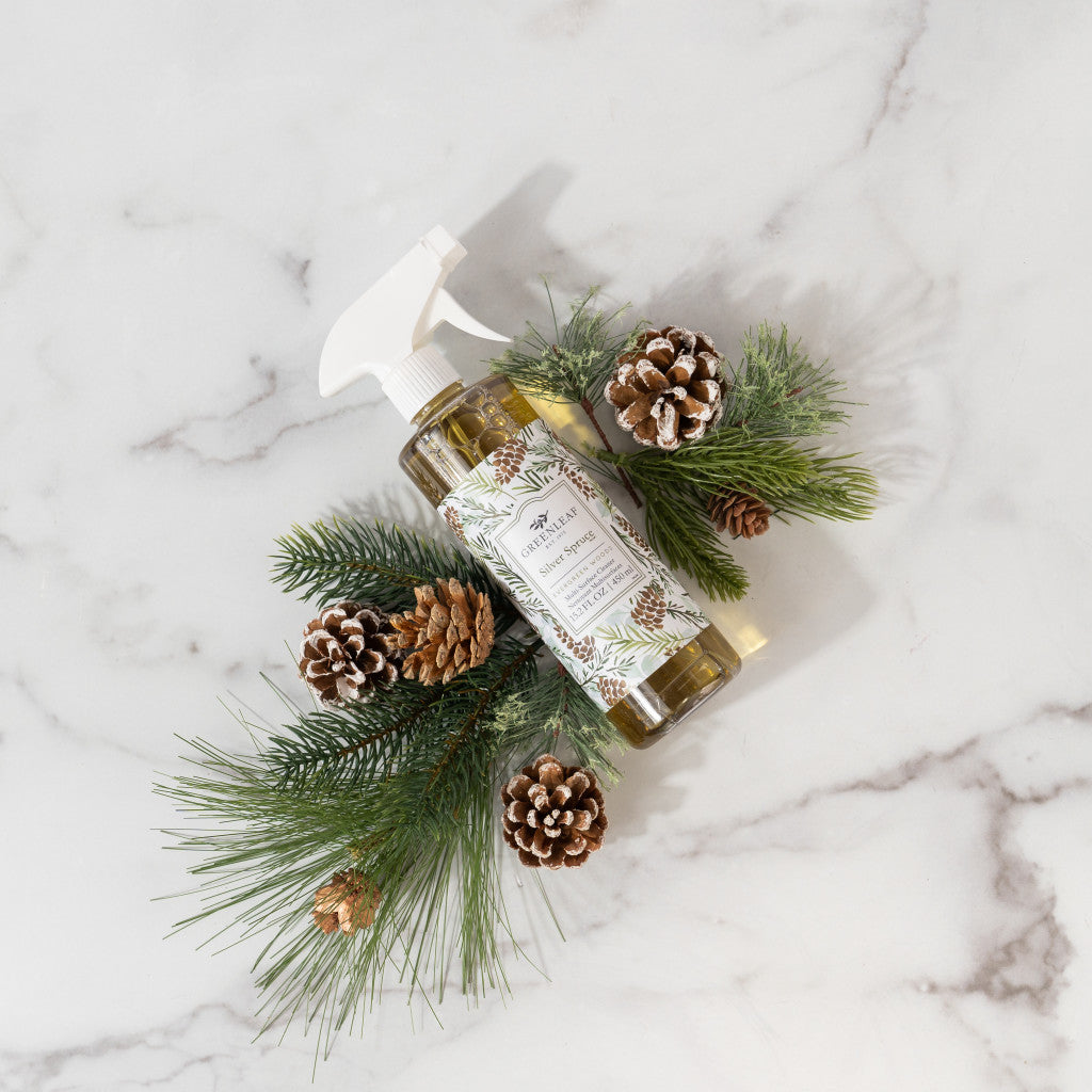 A spray bottle of Greenleaf Gifts’ Silver Spruce Multi-Surface Cleaner sits on marble, surrounded by pine branches and pinecones for a festive winter look—ideal for cleaning non-porous surfaces.