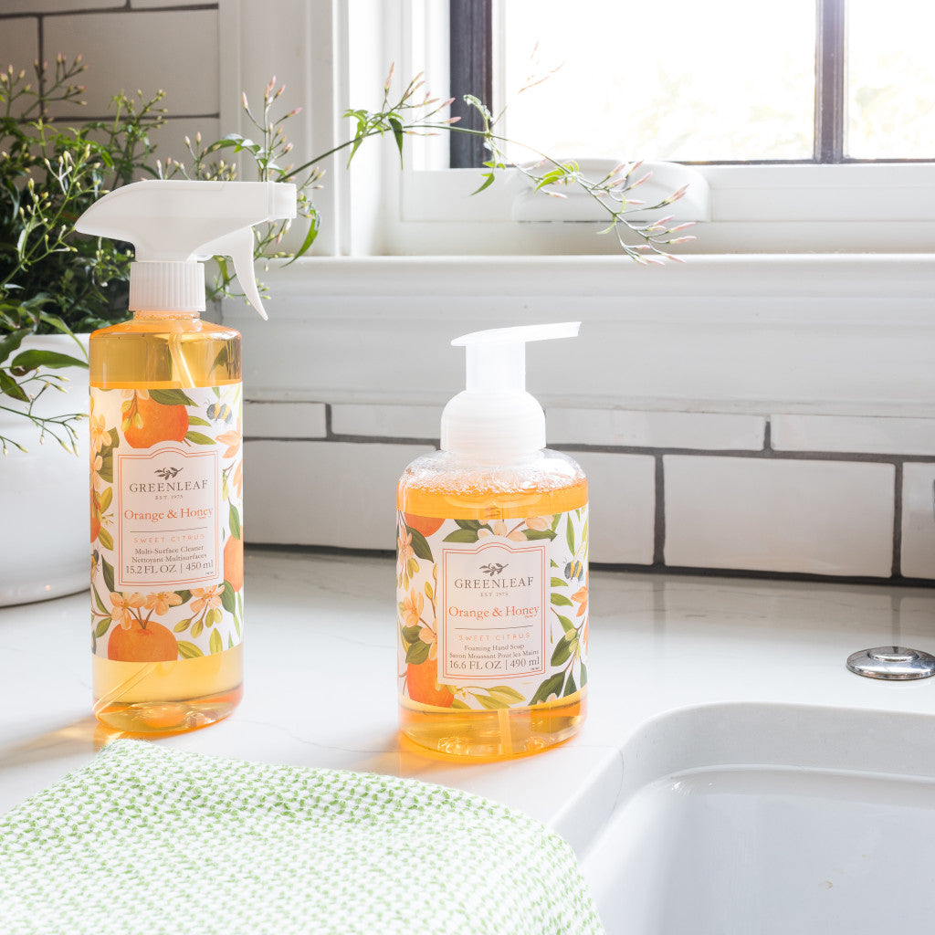 Two Greenleaf Gifts bottles with orange liquid—one Orange & Honey Multi-Surface Cleaner and one foaming hand soap—sit on a white kitchen counter near a sink and window, next to a green towel.