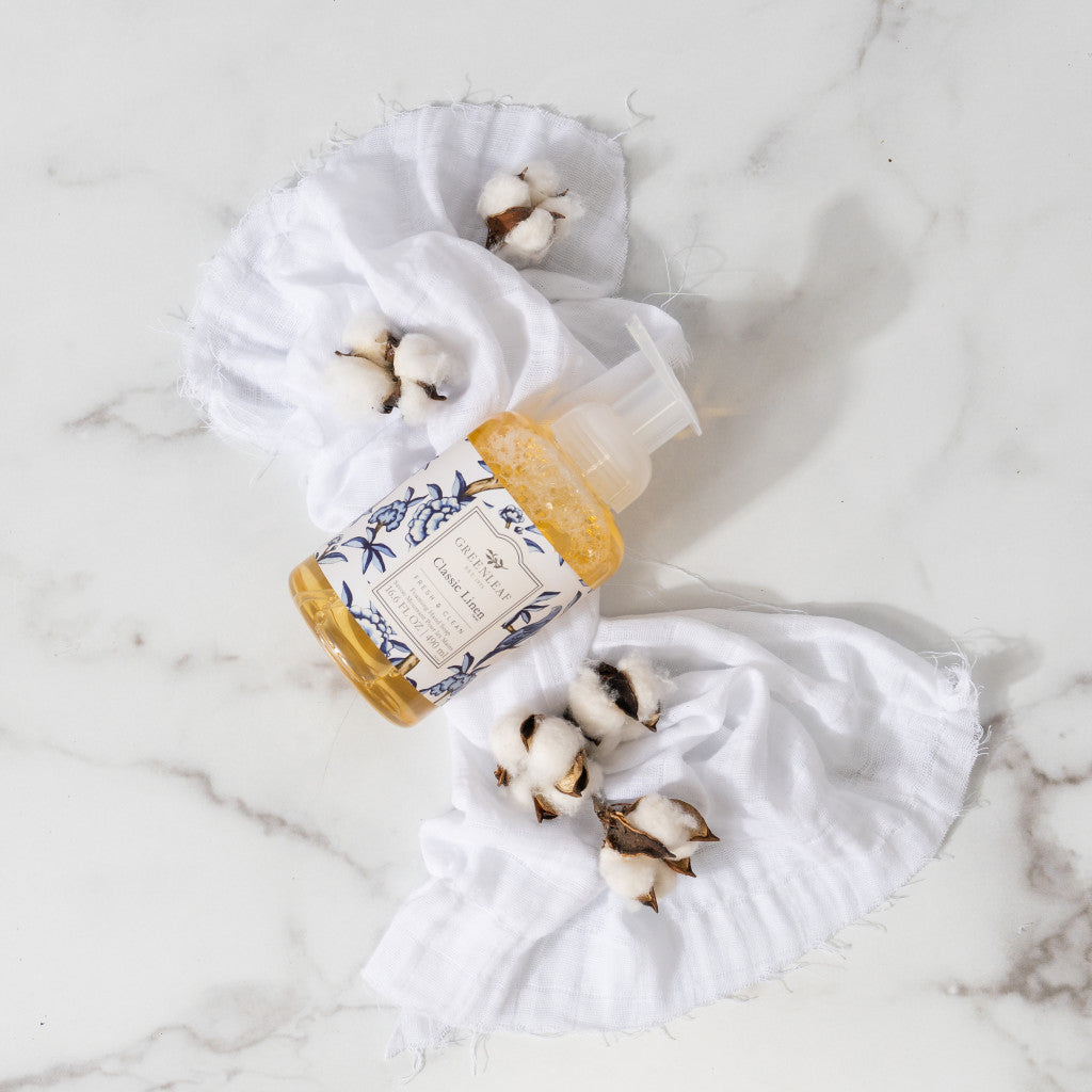 A bottle of Greenleaf Gifts Classic Linen Foaming Hand Soap with a decorative label sits on linen cloths among cotton branches, displayed on a white marble surface.