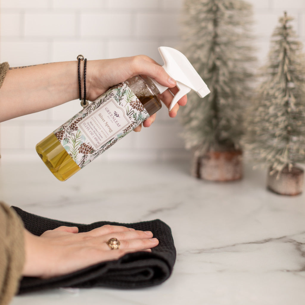 Someone sprays Greenleaf Gifts Silver Spruce Multi-Surface Cleaner onto a black cloth, with two frosted pine trees on a marble countertop—ideal for refreshing non-porous surfaces.