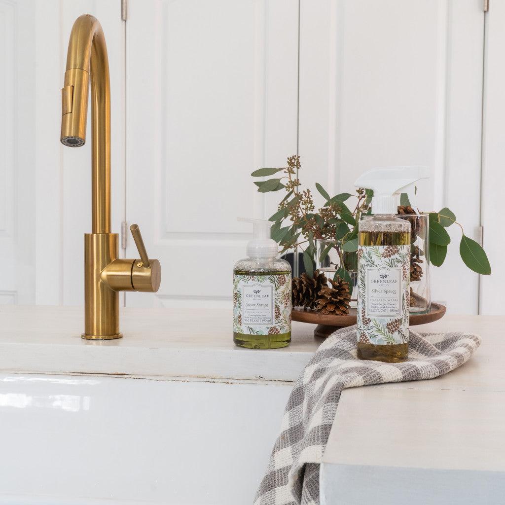 A kitchen sink with a gold faucet, a striped cloth on the counter, and two Greenleaf Gifts Silver Spruce Multi-Surface Cleaner bottles beside a small vase of greenery, set against white cabinets in soft natural light.