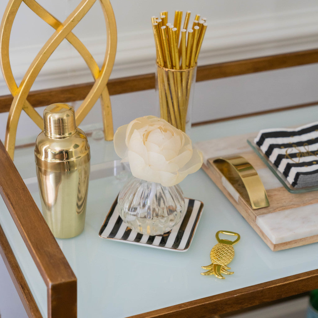 A stylish bar cart with gold accents features a gold cocktail shaker, golden straws in a holder, a striped coaster with a white flower, a pineapple bottle opener, and the Greenleaf Gifts Tuscan Vineyard Flower Diffuser for subtle fragrance.
