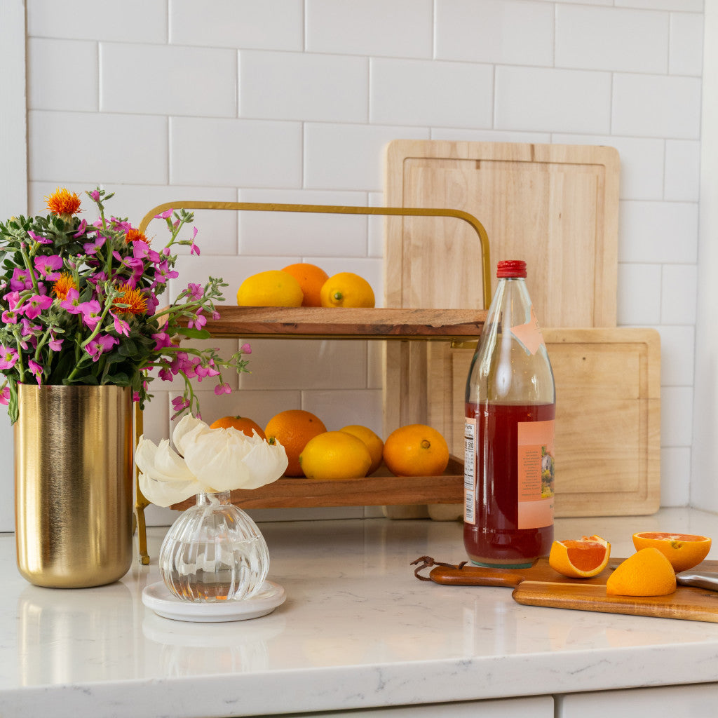 A kitchen counter with a two-tier wooden fruit stand of oranges, red juice, sliced oranges, a gold vase of pink flowers, the Greenleaf Gifts Tuscan Vineyard Flower Diffuser, and wooden cutting boards in the background.