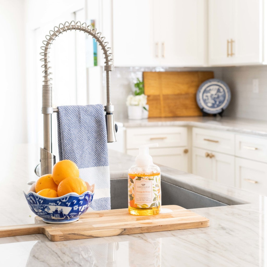 A modern kitchen features a marble countertop, a tall-faucet sink, blue-striped towel, bowl of lemons, and a Greenleaf Gifts Orange & Honey Foaming Hand Soap bottle. White cabinets and decor with a wooden cutting board are in the background.
