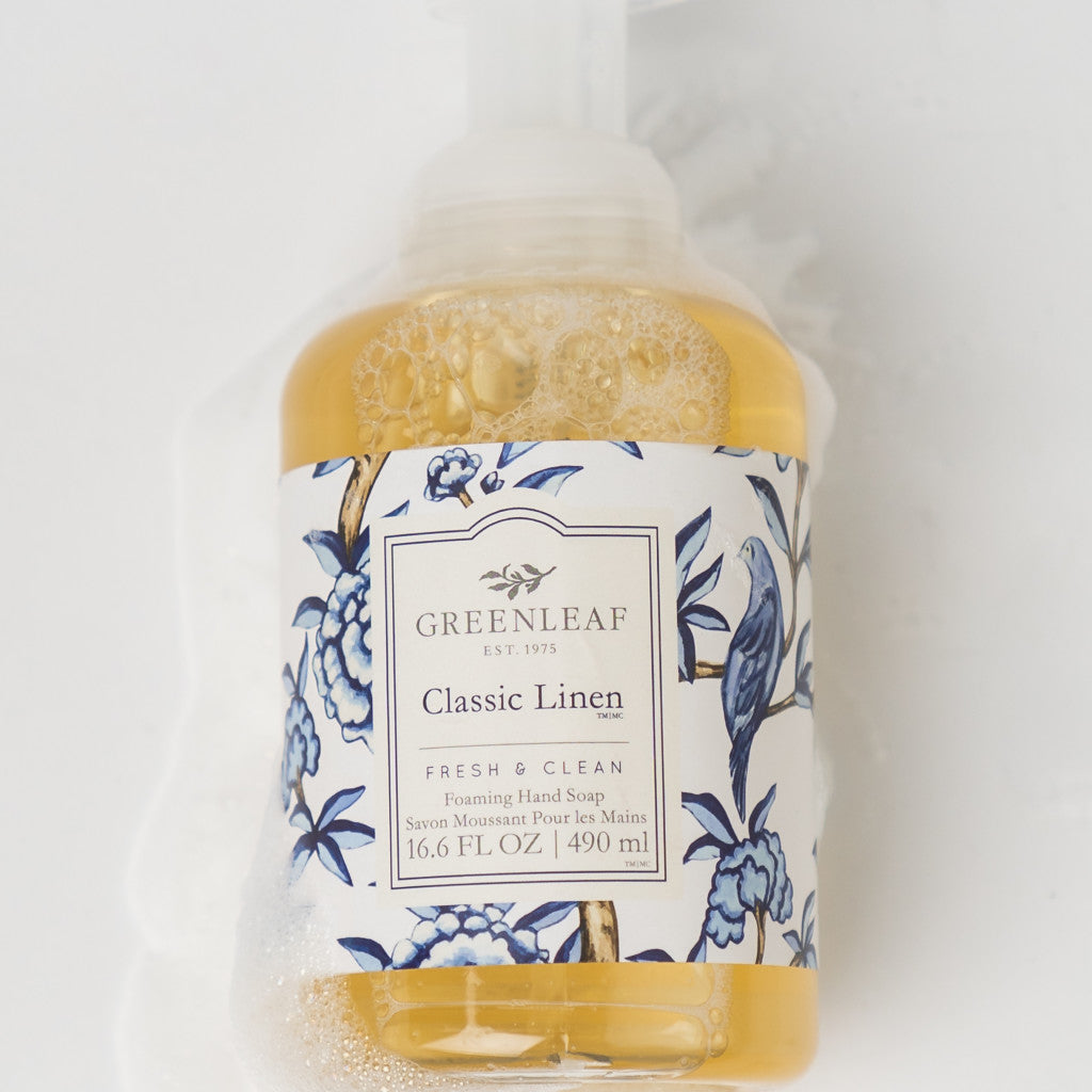 A bottle of Greenleaf Gifts Classic Linen Foaming Hand Soap, enriched with coconut oil, features a blue floral and bird label with soap suds on a white background.