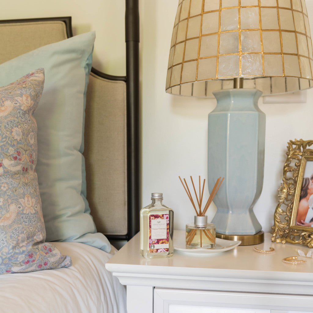 A cozy bedside scene with pillows, a blue ceramic lamp, a bottle, Greenleaf Gifts Tuscan Vineyard Diffuser Oil Refill, jewelry, and a framed photo on a white nightstand.