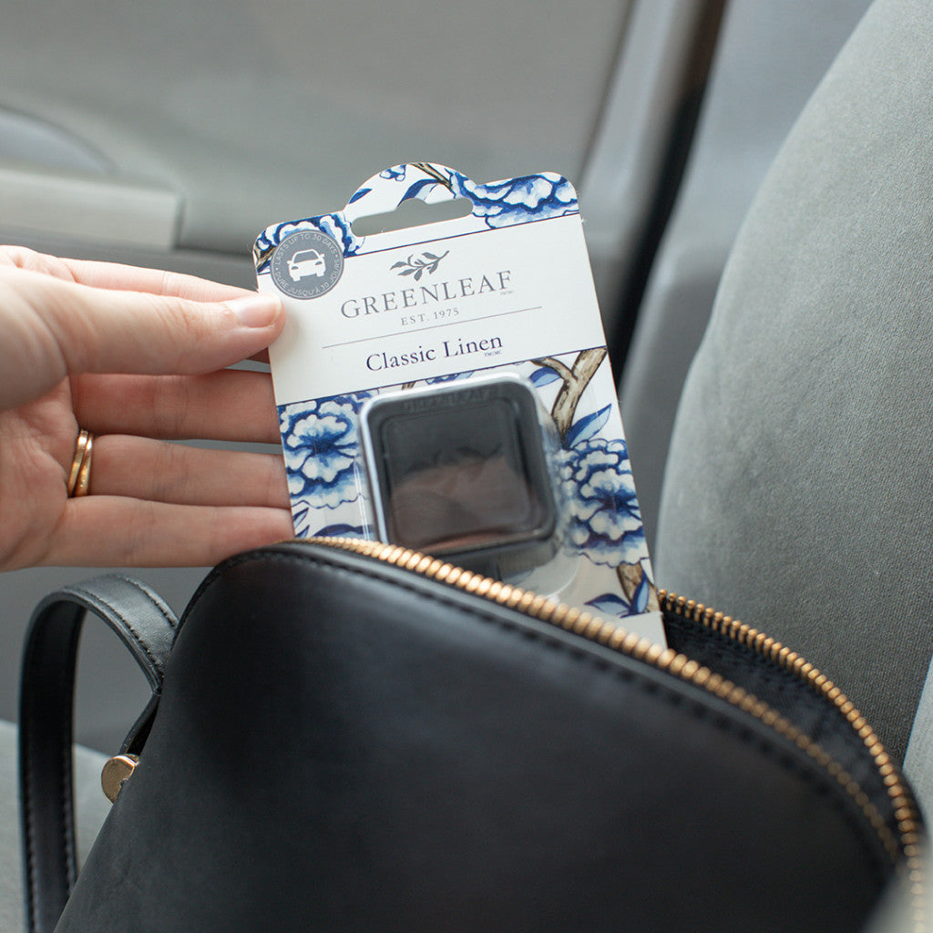 A hand holds a Greenleaf Gifts Classic Linen Auto Vent Clip car air freshener package above an open black purse on a gray car seat.