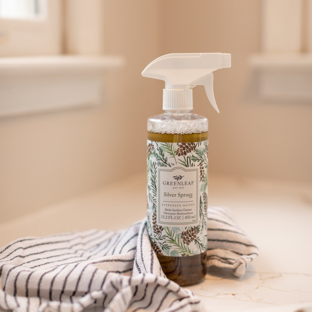 A bottle of Greenleaf Gifts Silver Spruce Multi-Surface Cleaner, featuring a pinecone and pine needles design label, rests on a striped towel atop a light counter—perfect for refreshing non-porous surfaces.