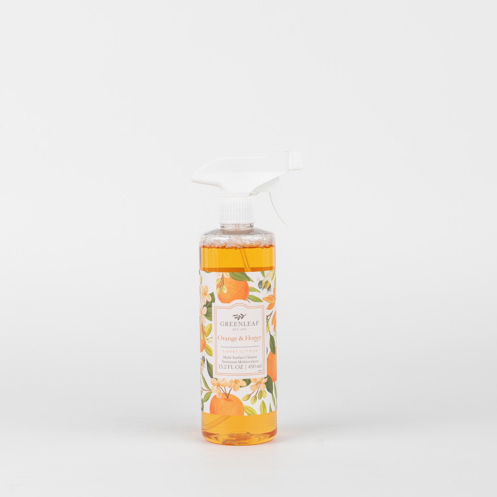 A clear Greenleaf Gifts Orange & Honey Multi-Surface Cleaner spray bottle, filled with orange liquid and featuring orange and leaf illustrations, is shown against a plain white background.