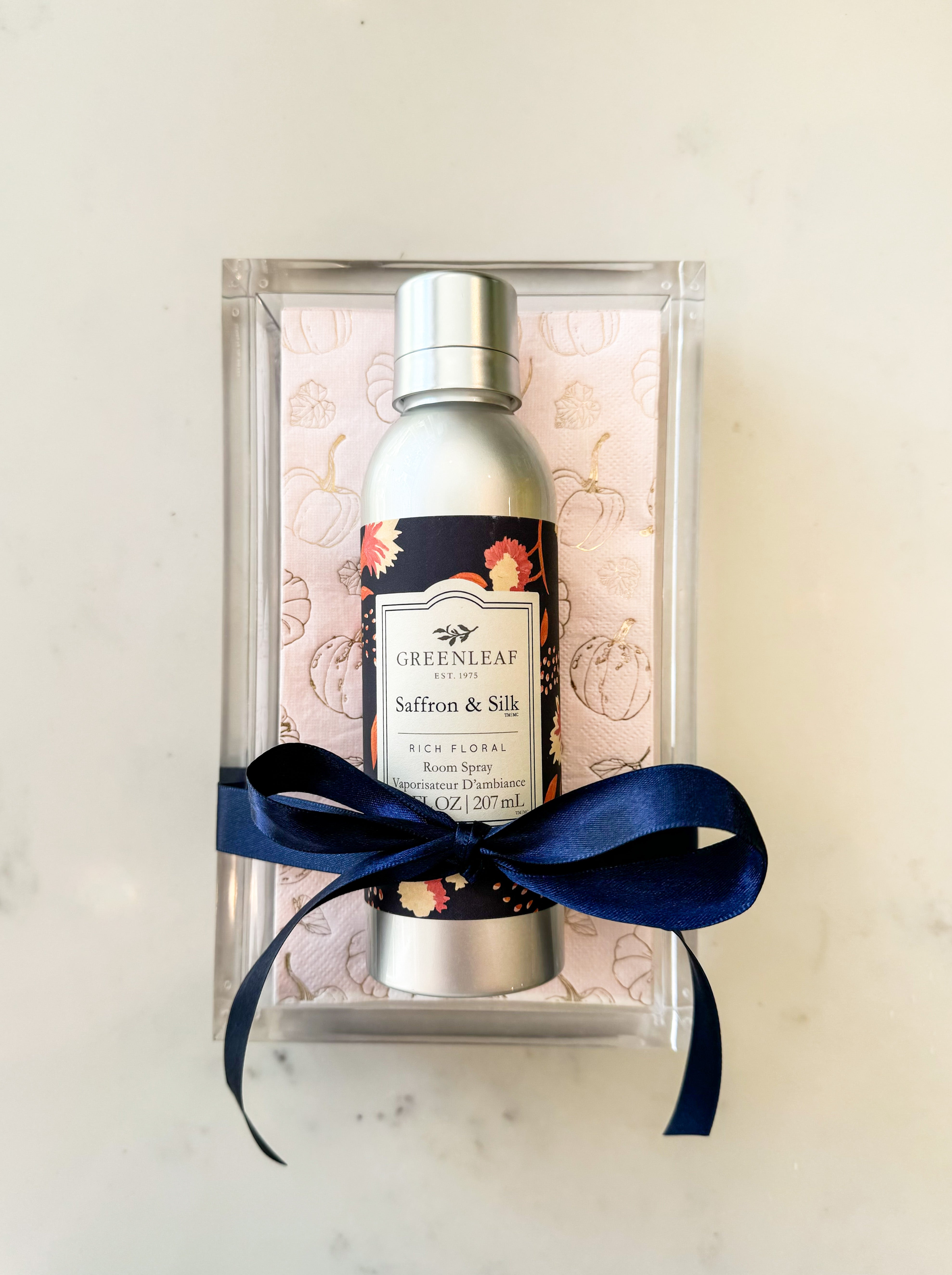 A silver bottle of Greenleaf Gifts Saffron & Silk Room Spray sits on a pink patterned cloth in a clear box, tied with a dark blue ribbon.