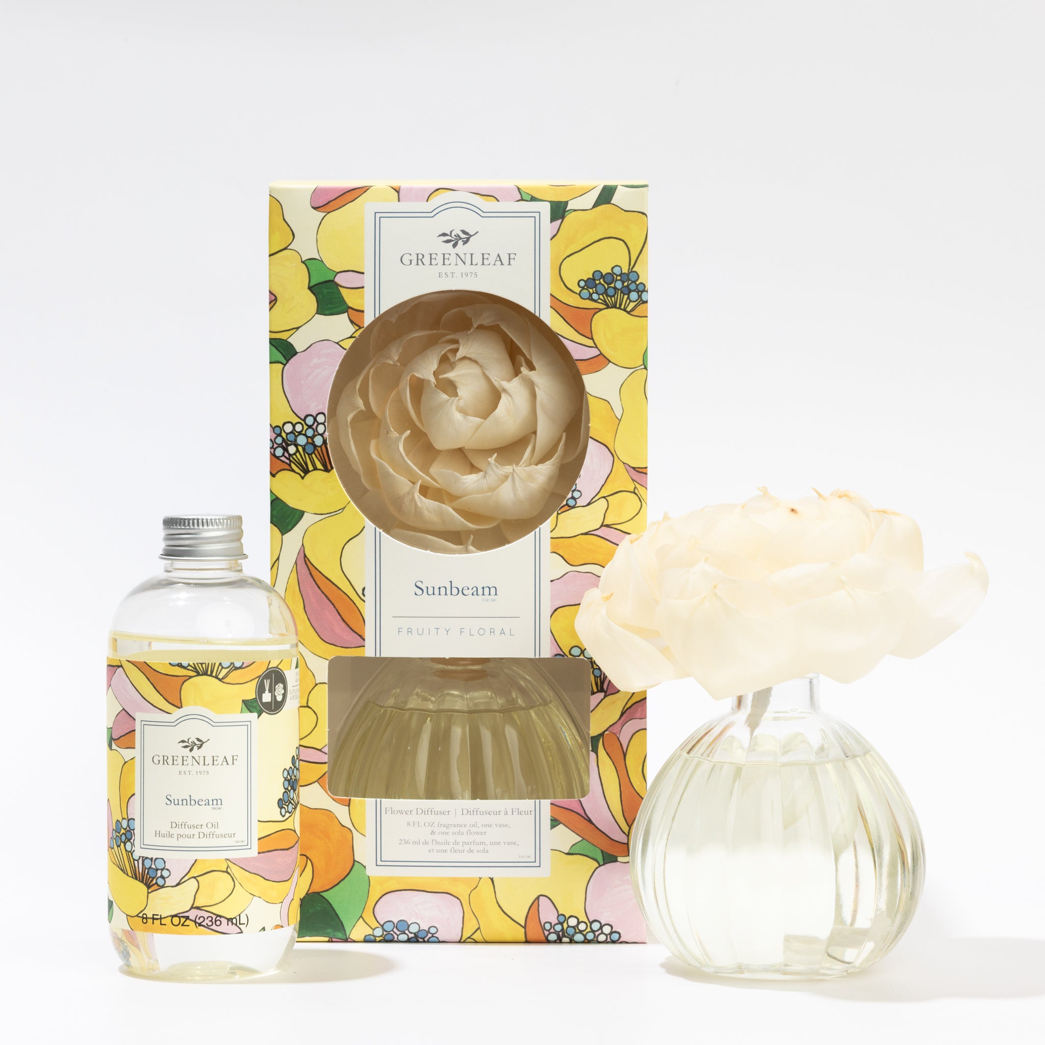 A Sunbeam Flower Diffuser & Fragrance Oil Refill by Fast Bundle, in a colorful floral box with yellow, pink, and green accents, sits beside a clear vase holding a white flower diffuser.