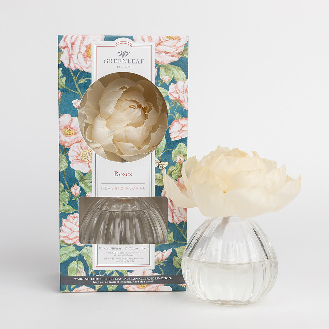 The Greenleaf Gifts Roses Flower Diffuser features a sola paper flower atop a clear glass bottle filled with fragrance oil, next to its rose-patterned box, ready to infuse your space with a classic floral scent.