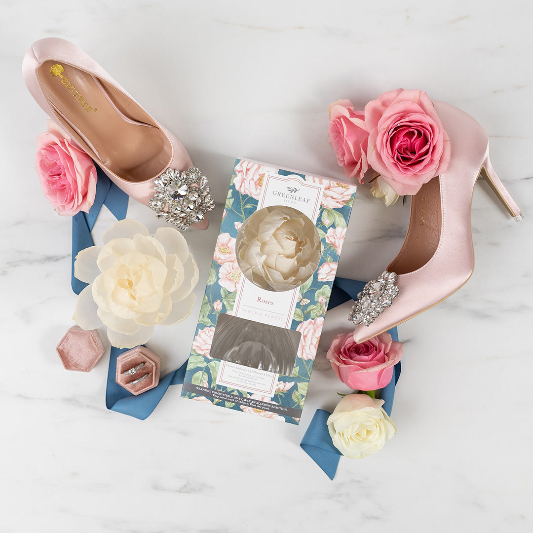 A flat lay features pink high-heeled shoes with jeweled accents, surrounded by pink and white roses, a blue ribbon, a Greenleaf Gifts Roses Flower Diffuser box with a floral pattern, and a pink ring box on marble.