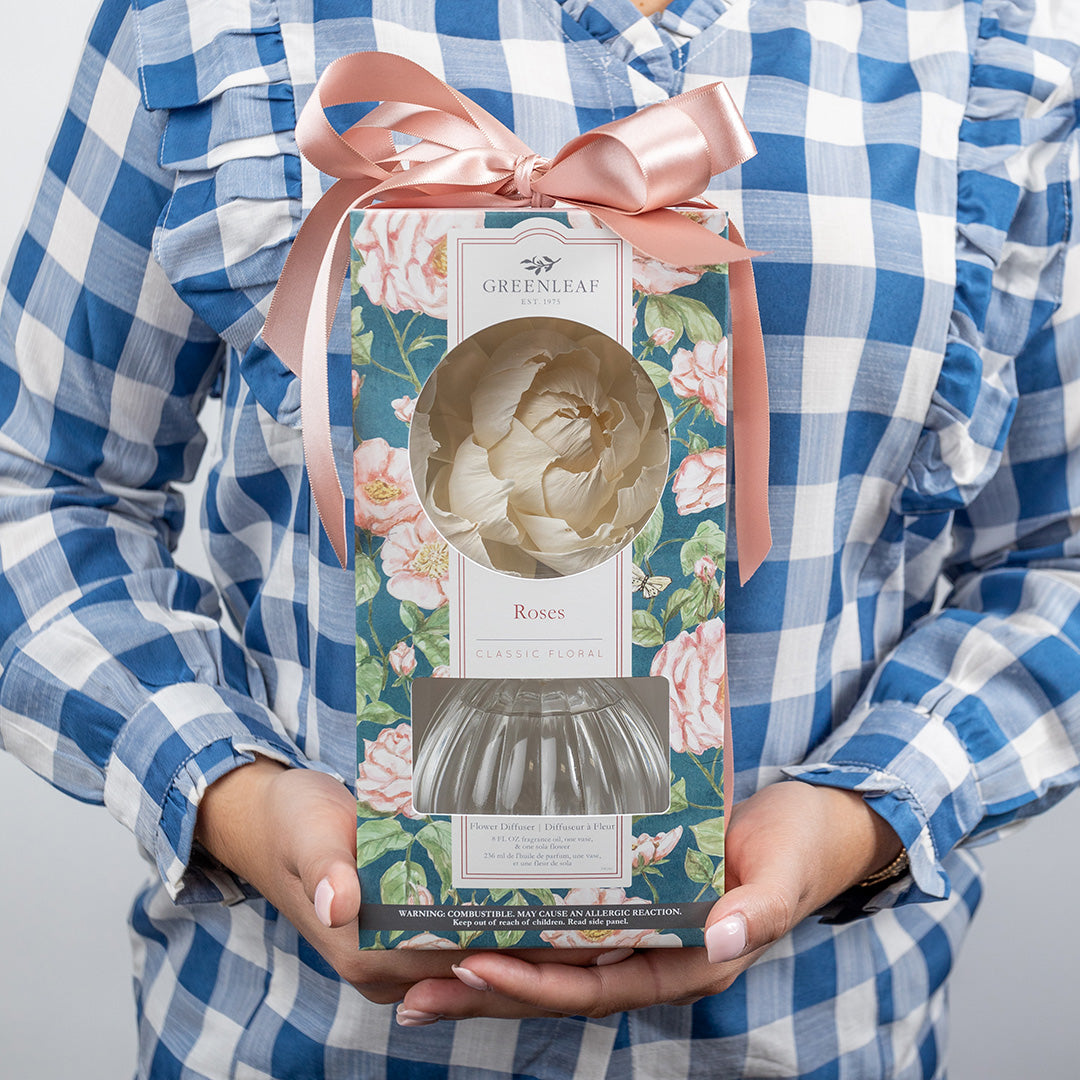 A person in a blue and white plaid shirt holds a Greenleaf Gifts Roses Flower Diffuser box with a pink ribbon. The box displays rose-scented décor, fragrance oil, and a sola paper flower through its window.