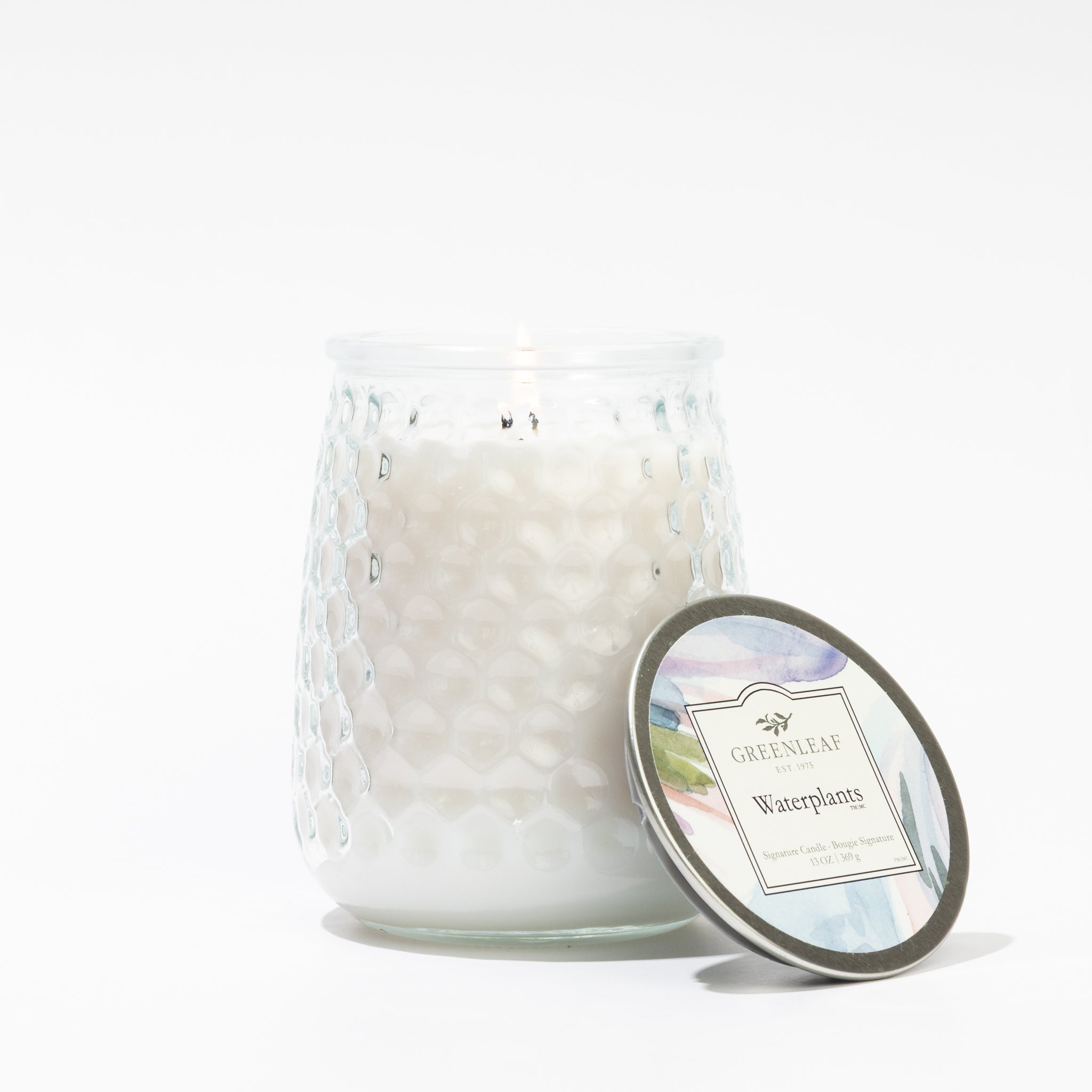 A lit Greenleaf Gifts Waterplants Signature Candle in a textured glass jar sits beside its decorative lid, which features "Greenleaf Waterplants" on a white background.