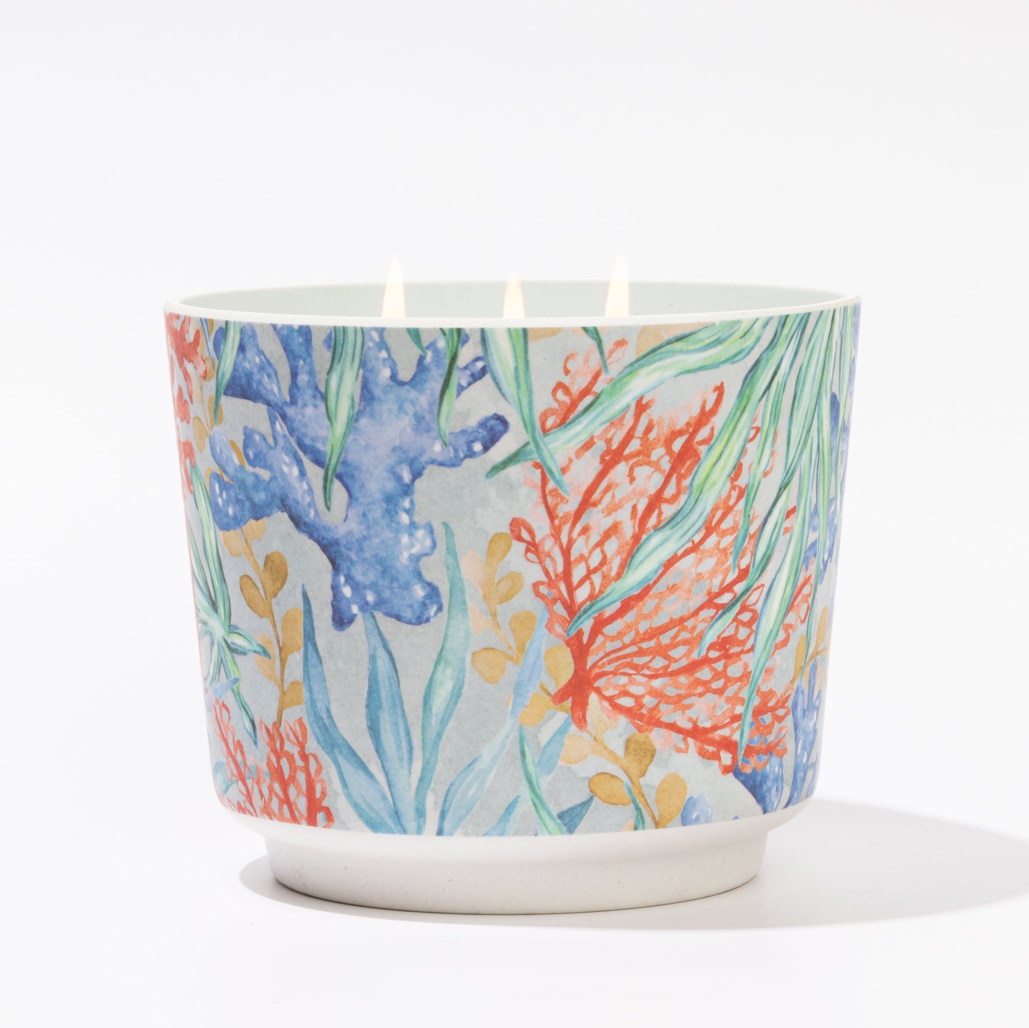 The Greenleaf Gifts Seaspray Patterned 3 Wick Candle features a ceramic holder adorned with blue, red, and green coral reef designs. Three candles burn brightly in the holder, displayed on a white surface with a white background.