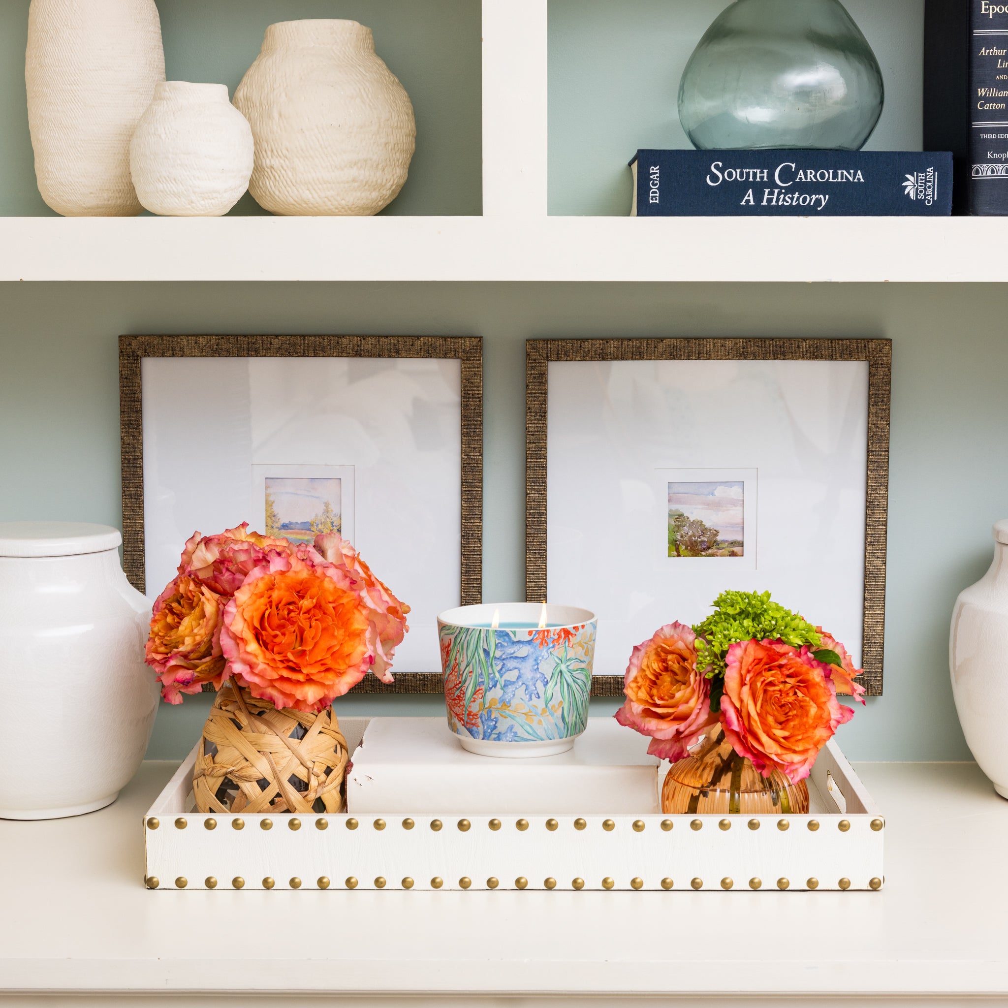 A white tray with the Greenleaf Gifts Seaspray Patterned 3 Wick Candle and two small vases of orange flowers sits on a shelf, surrounded by white pottery, framed art, and books on the upper shelves.