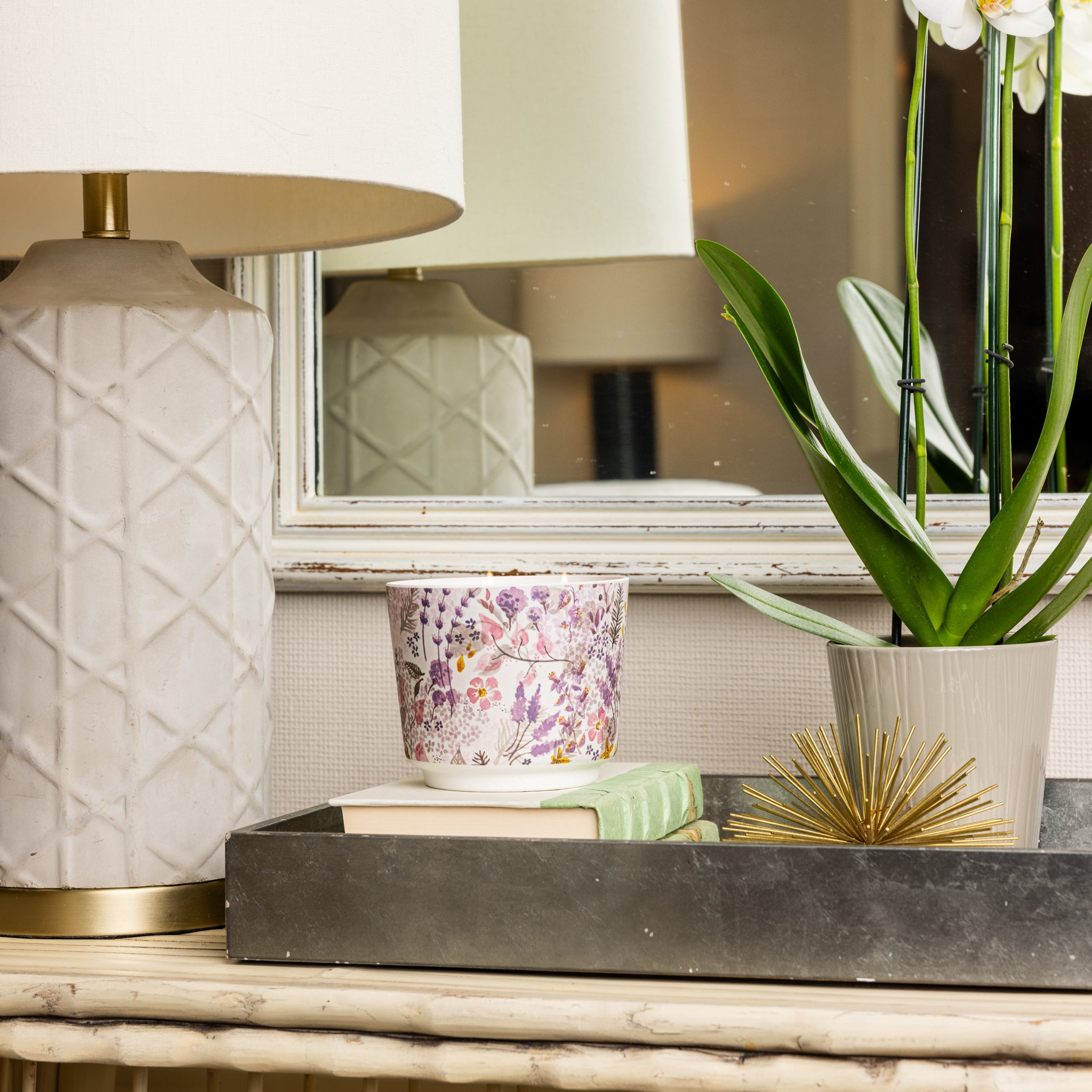 A decorative table holds a white lamp, a floral-patterned mug, a potted orchid, books, and the Greenleaf Gifts Lavender Patterned 3 Wick Candle, all arranged on a tray in front of a mirror.