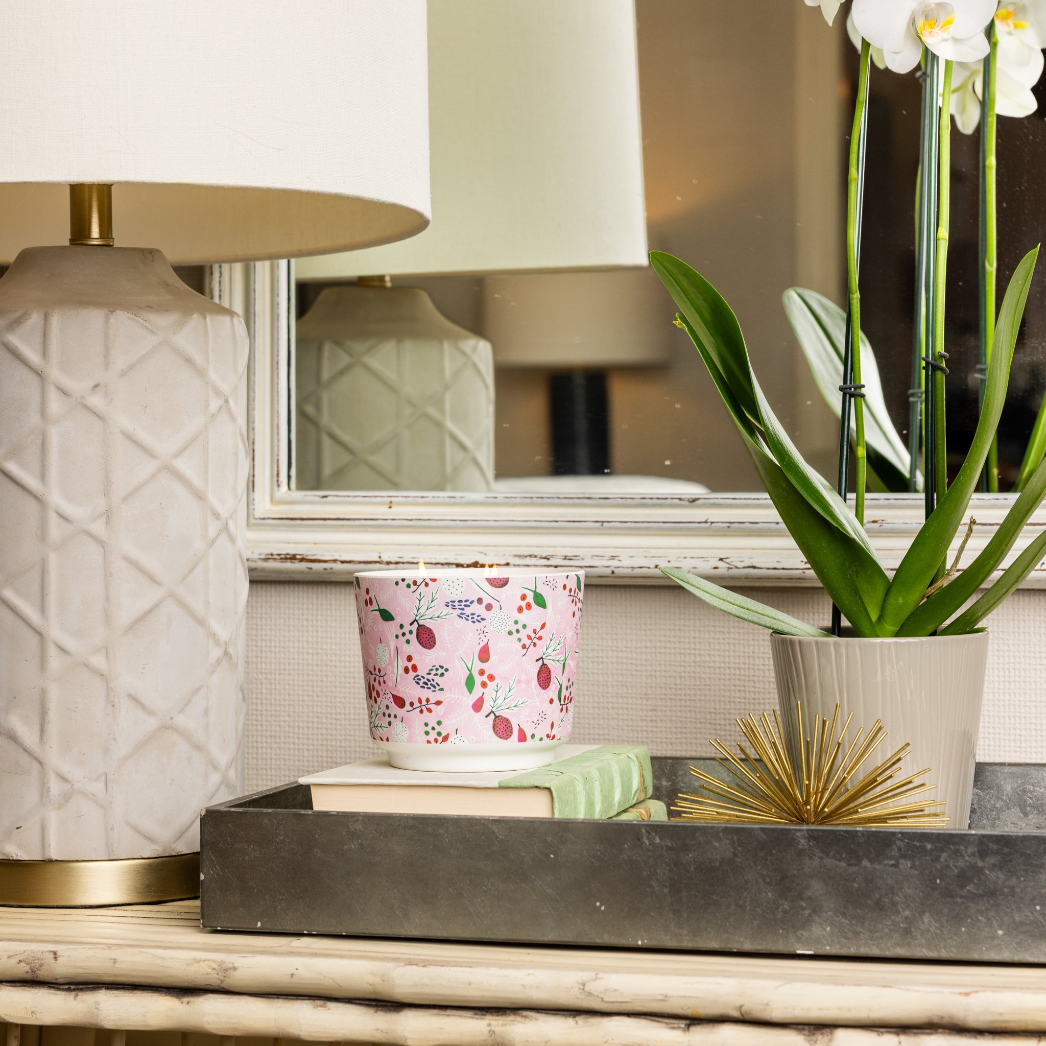 A decorative table displays a textured lamp, a floral mug, stacked books, the Brambleberry Patterned 3 Wick Candle by Greenleaf Gifts, a gold spiky ornament, and a potted orchid on a metal tray before a mirror.