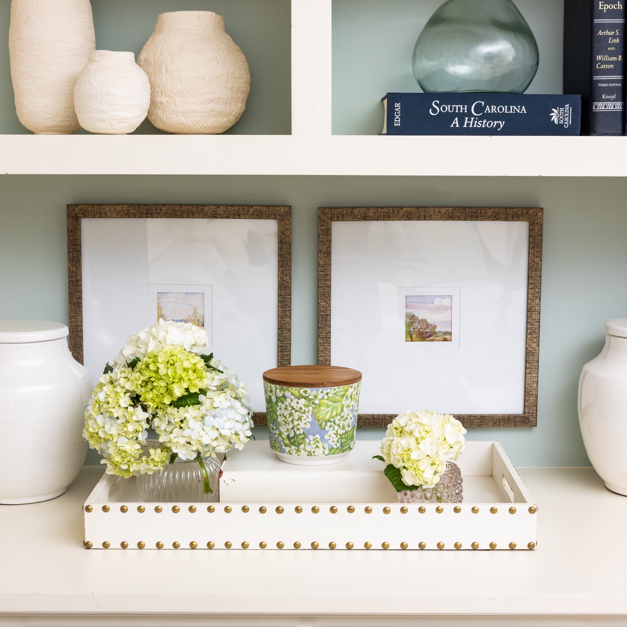 A white shelf displays two framed pictures, a book, textured vases, a glass vase, and a tray with a floral container and small bouquets of white and green hydrangeas beside Greenleaf Gifts’ Hydrangea Patterned 3 Wick Candle.