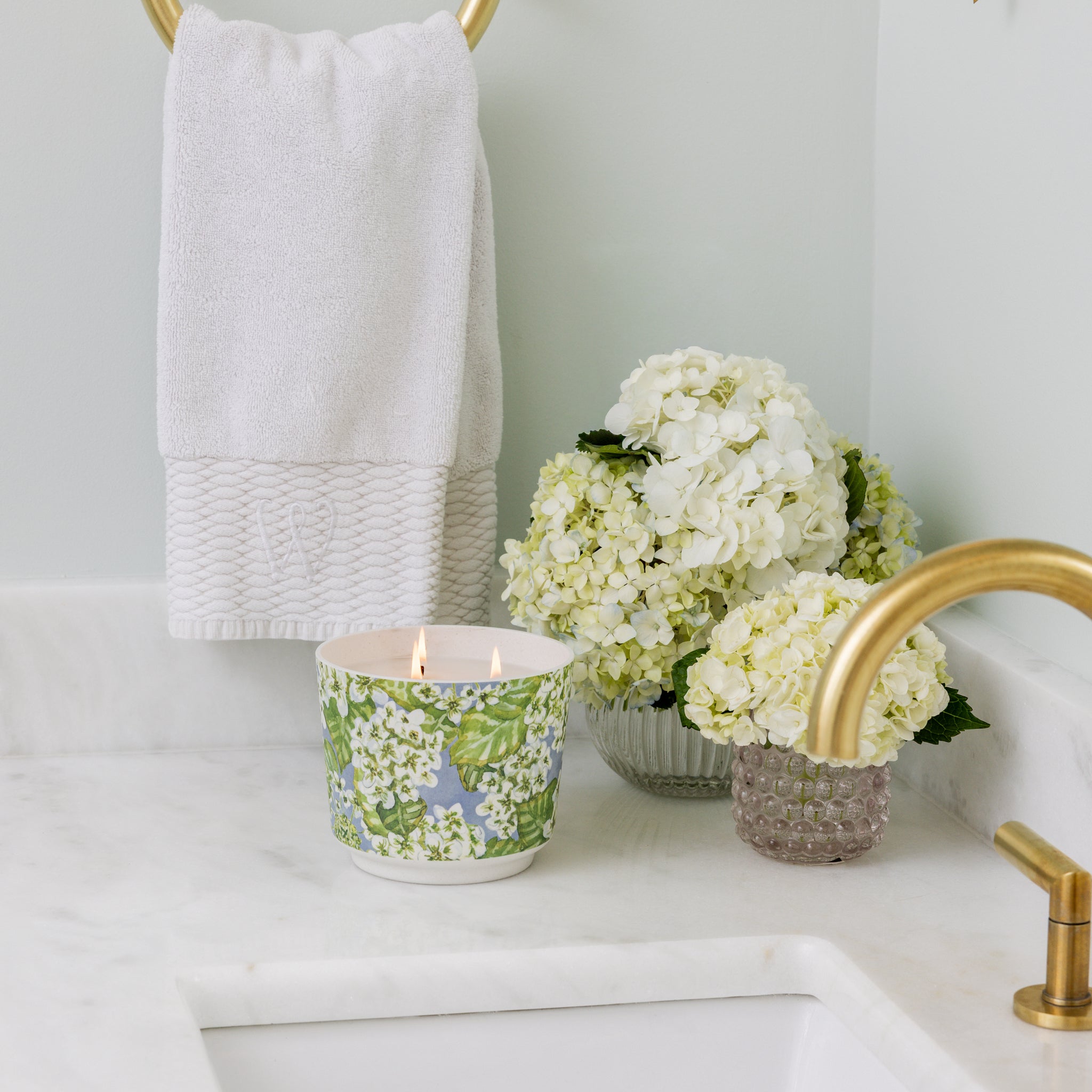 A bathroom countertop features a gold faucet, a white towel on a gold ring, and the Hydrangea Patterned 3 Wick Candle by Greenleaf Gifts, filling the space with hydrangea fragrance. White hydrangeas in a glass vase add an elegant, fresh touch.