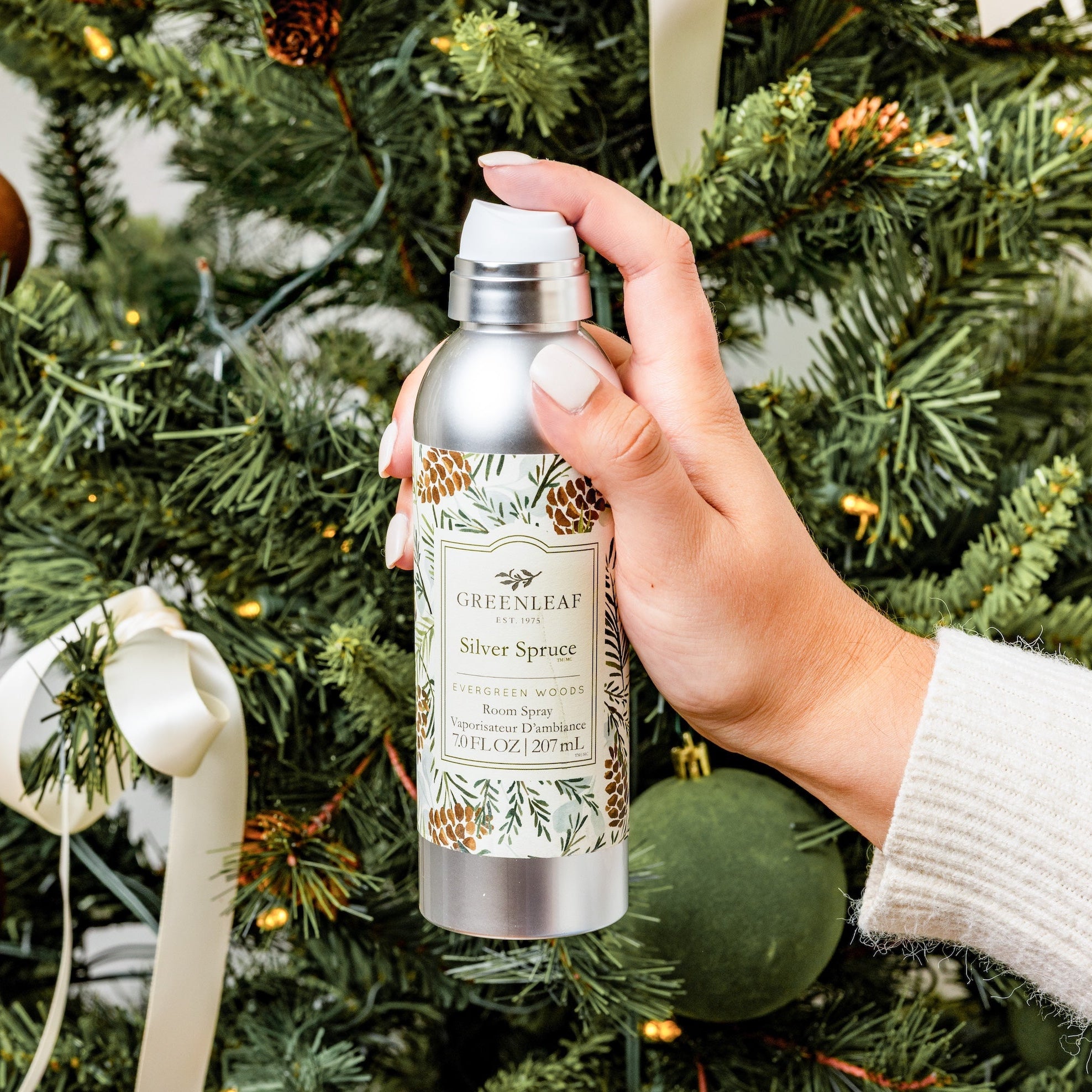 Hand holding a Greenleaf Silver Spruce Room Spray bottle in front of a decorated Christmas tree.