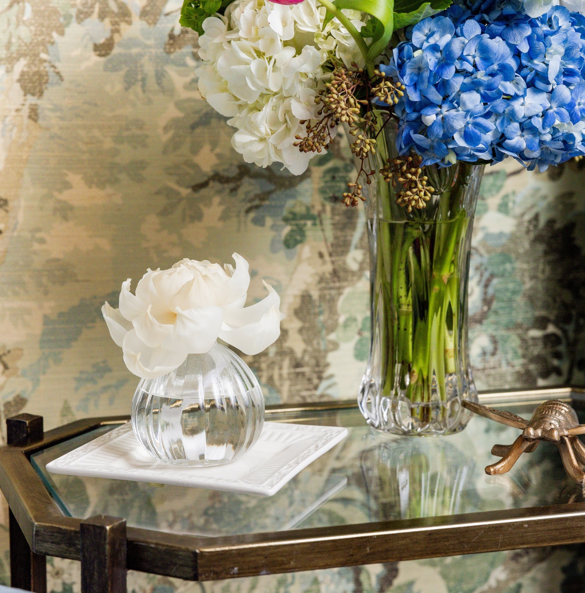 The Peony Bloom Flower Diffuser by Greenleaf Gifts sits on a napkin on a glass table beside a larger vase of white, blue, and green flowers, creating an elegant botanical display against patterned wallpaper.