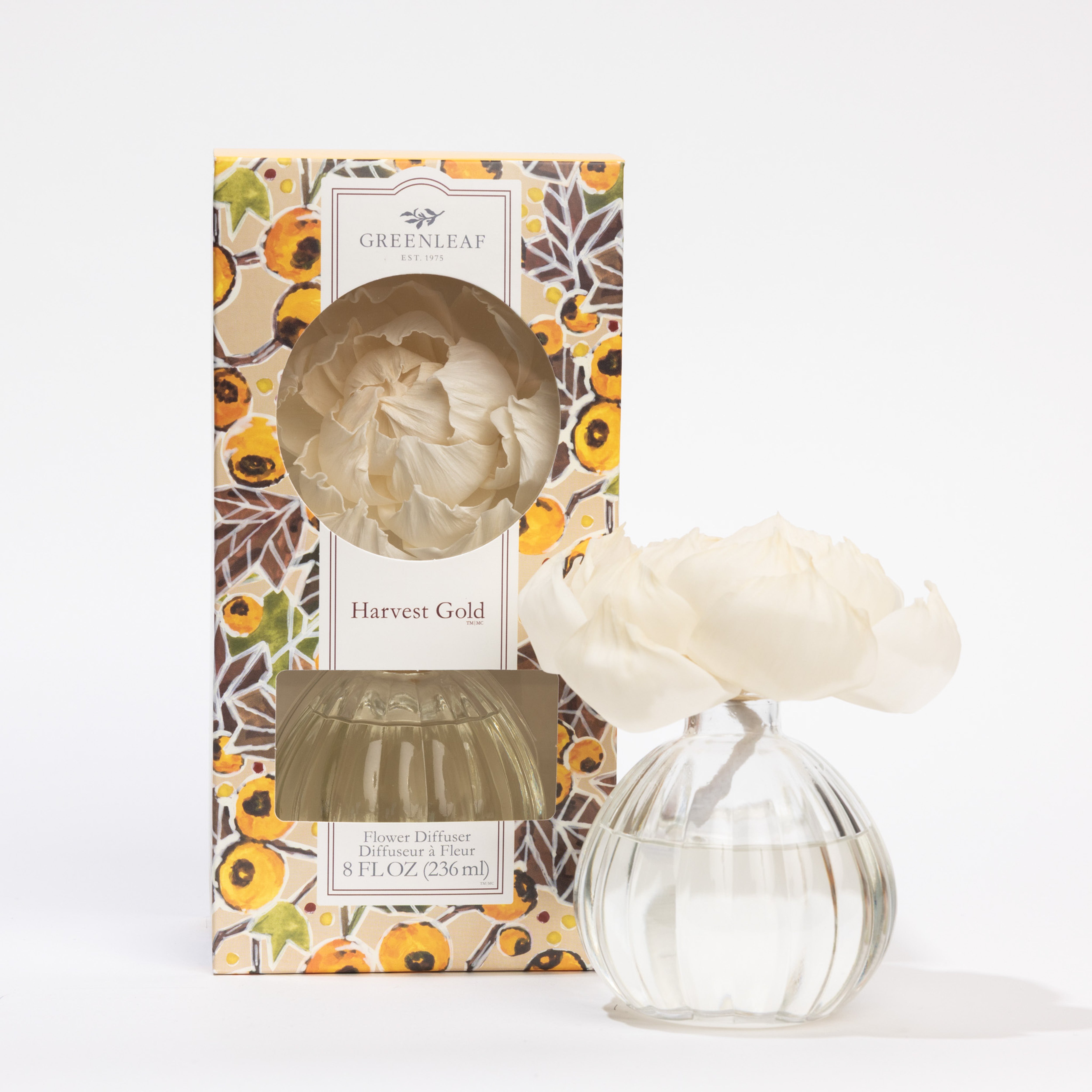 A clear glass Harvest Gold Flower Diffuser by Greenleaf Gifts, topped with a white sola flower, sits next to its floral-patterned box labeled "Greenleaf Gifts." The box features a window showing the fragrant diffuser inside.