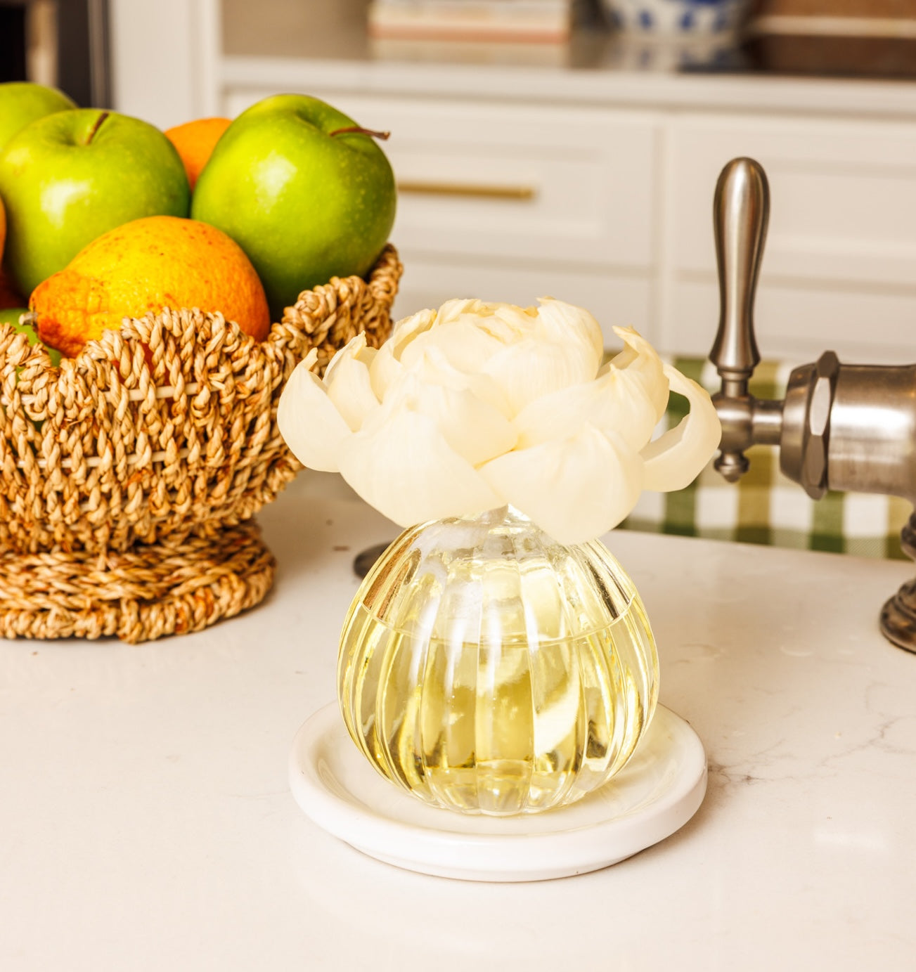 A Greenleaf Gifts Orange & Honey Flower Diffuser, featuring clear glass filled with pale fragrance oil and topped with a white sola paper flower, rests on a marble tray beside a woven fruit basket of apples and pears on the kitchen counter.