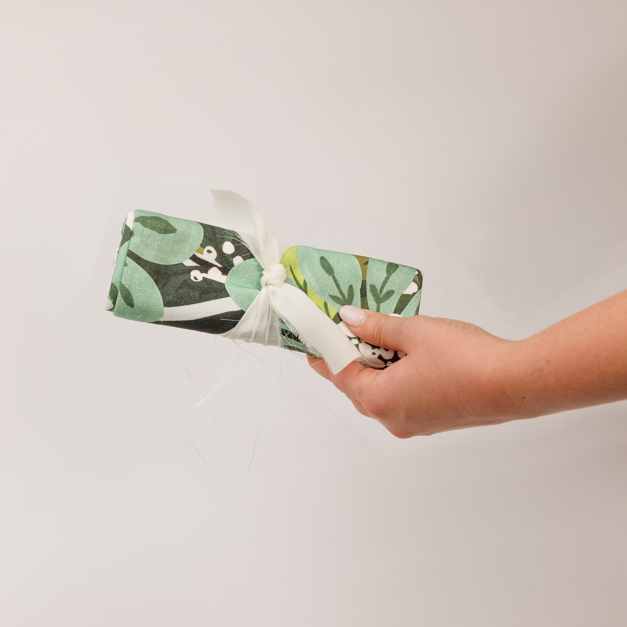 A hand holds a rectangular gift wrapped in green and white floral paper with a white ribbon, against a plain white background—an ideal way to present the Shimmering Snowberry Printed Tea Towel by Greenleaf Gifts this holiday season.