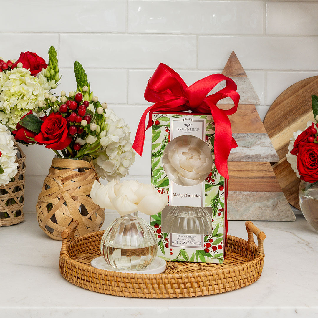 The Merry Memories Flower Diffuser by Greenleaf Gifts rests on a wicker tray beside its holly-adorned, red-bowed box. White subway tiles, a wooden tree, and vases of red and white flowers complete this festive, fragrant display.